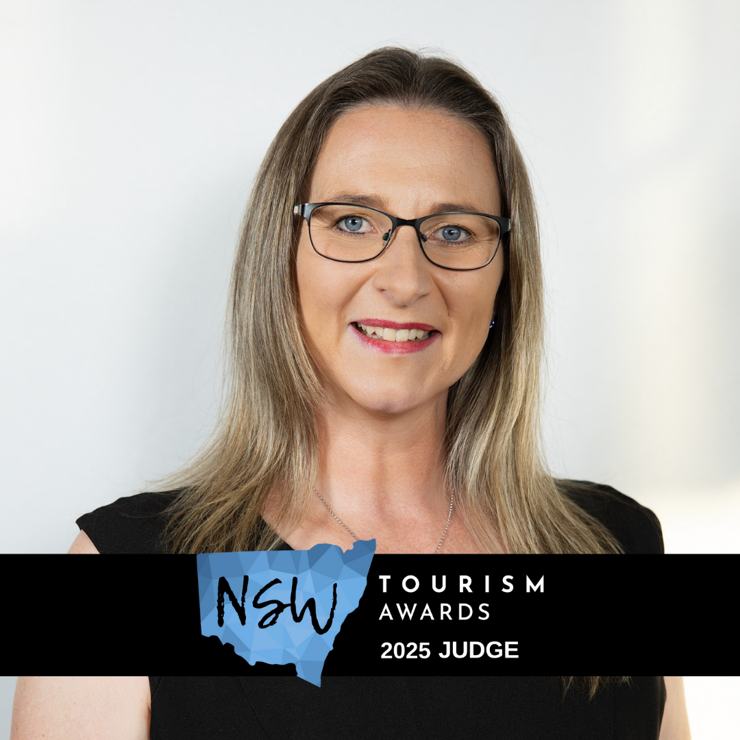 Portrait of a woman with shoulder-length blonde hair wearing glasses and a black sleeveless top. She is smiling and standing in front of a plain background. The overlay text reads "NSW Tourism Awards 2025 Judge" with a blue geographic shape of New South Wales.