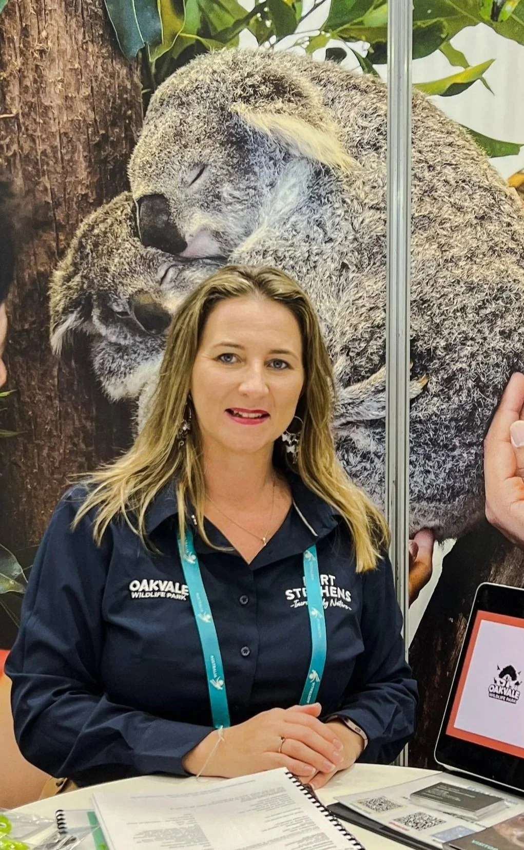 A woman with blonde hair wearing a dark blue shirt with 'OAKVALE WILDLIFE PARK' and 'Port Stephens' printed on it, sitting at a table with papers, with a large background poster of two koalas cuddling.