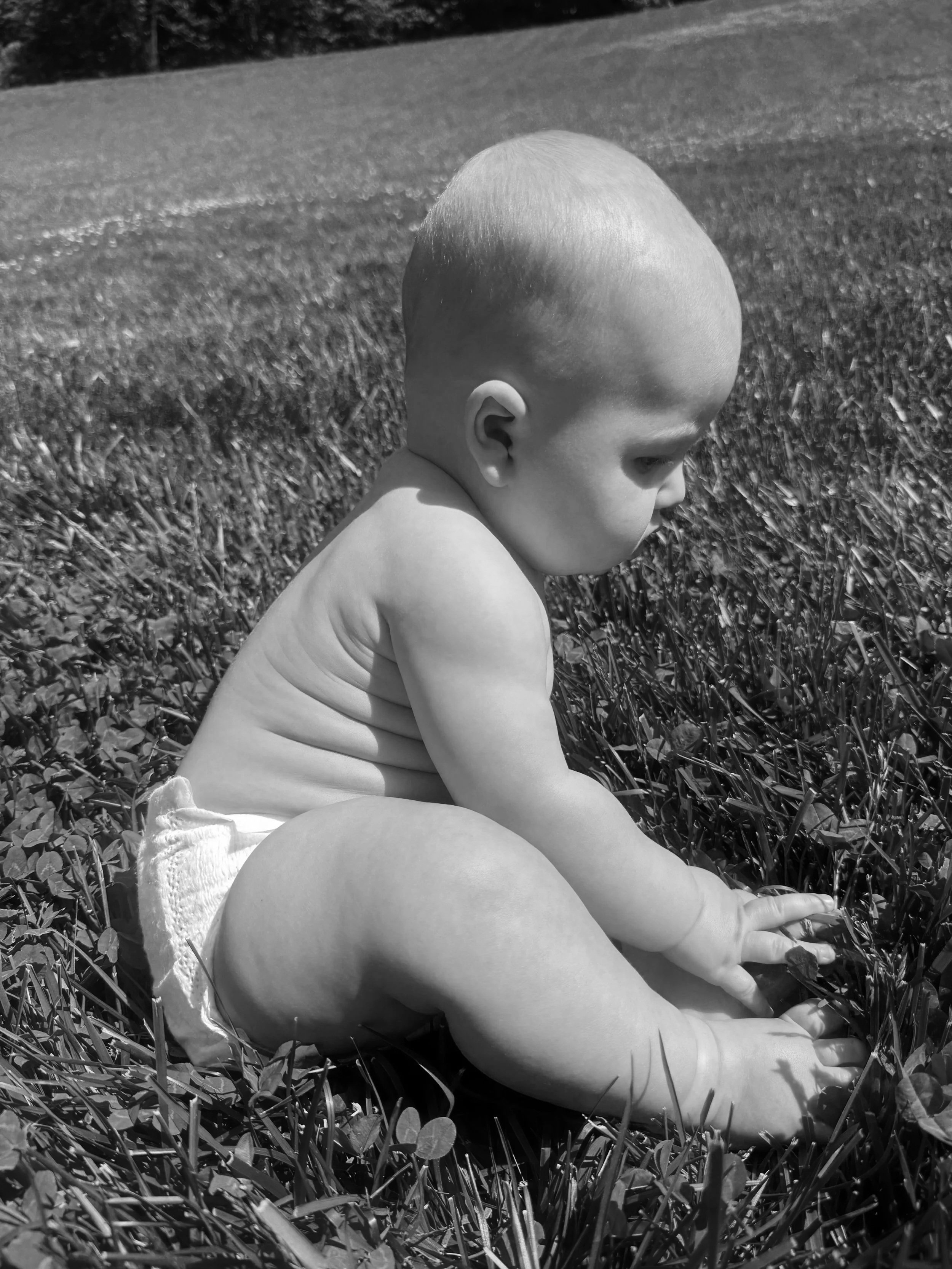 A young child with no shirt and a diaper sitting on grass, leaning forward and touching the ground, with a blurred background of grass and trees.