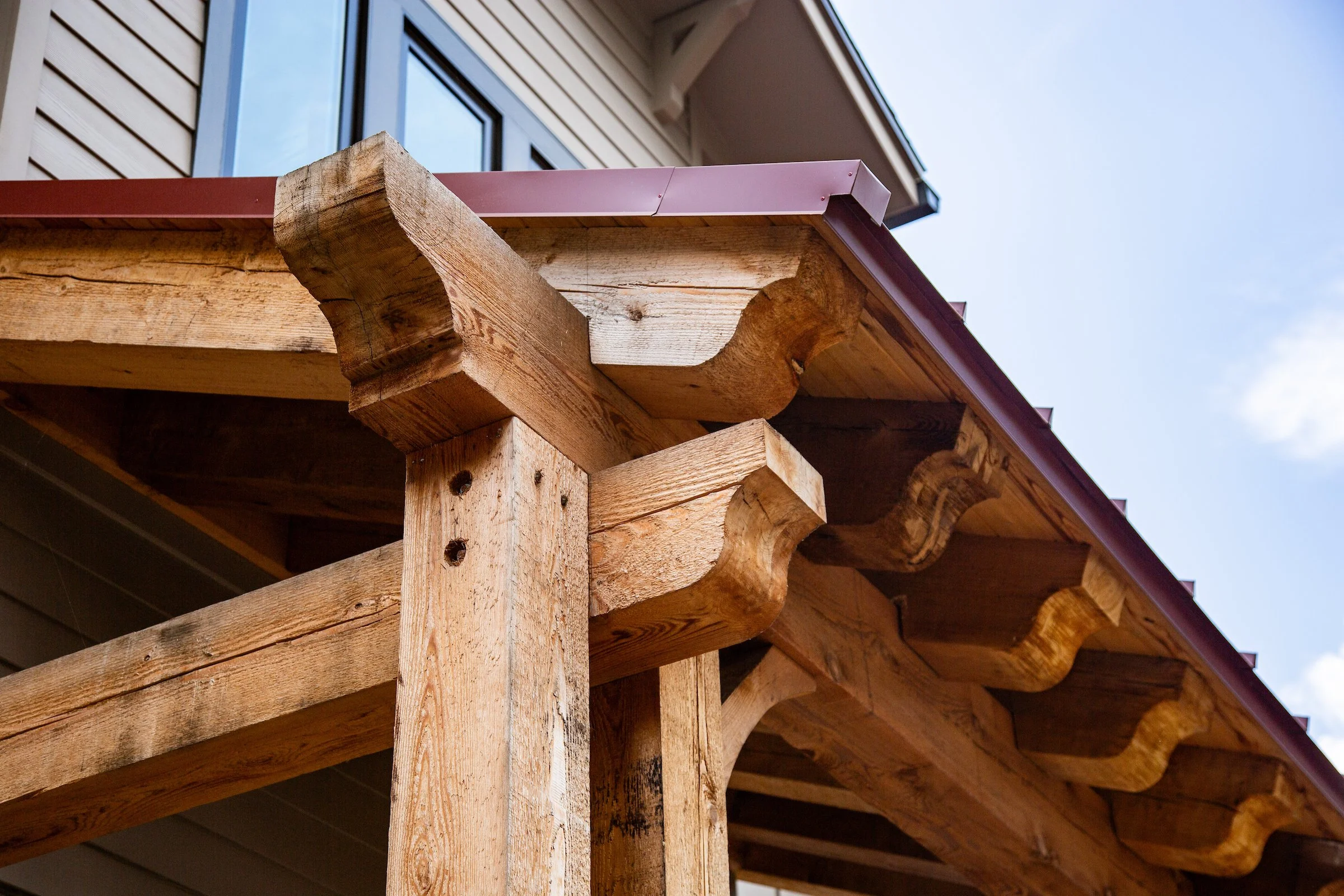 Close-up of wooden structural beams and supports of a building's roof with a metal gutter above in a partly cloudy sky.