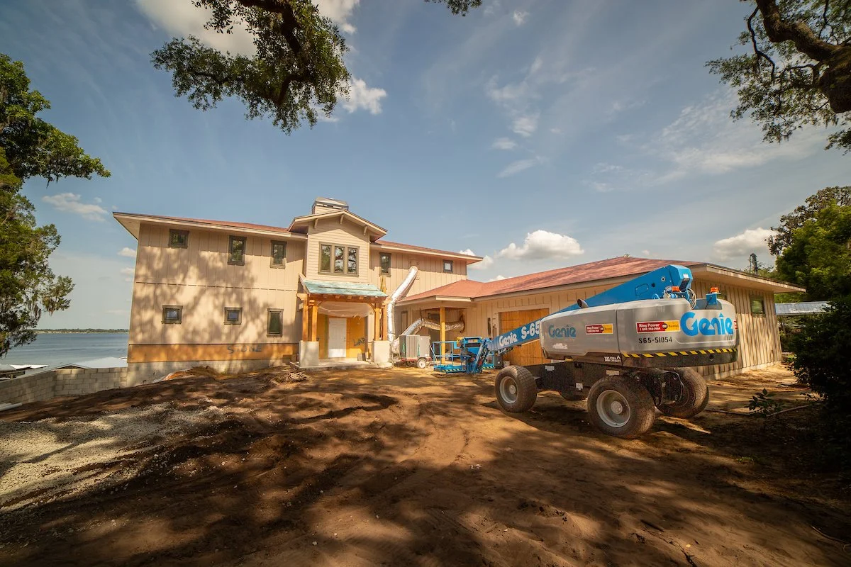 A house under construction near a body of water, with construction equipment like a lift and ductwork visible on the dirt yard.