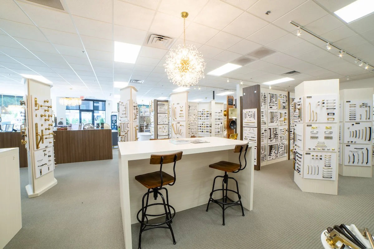Interior of a store showcasing various door handles and knobs displayed on white panels, with a central white table and two wooden chairs in the foreground.