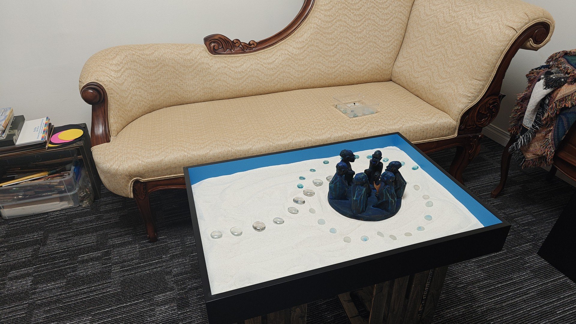 A living room with a beige vintage-style sofa, a coffee table with a sand-colored surface, and a side table with books and CDs. The coffee table features a small sculpture of four figures surrounded by glass pebbles on a white surface.