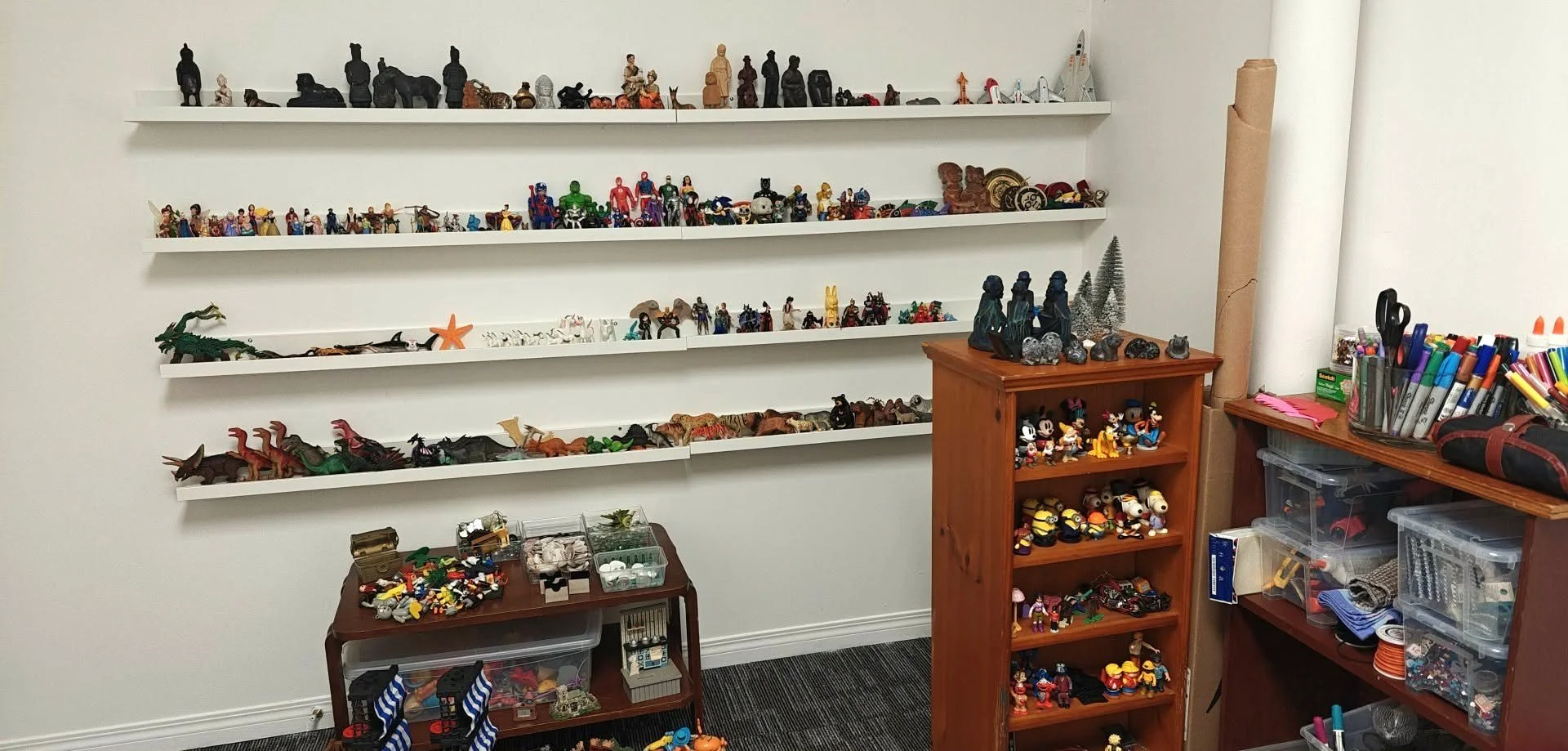 Room with white wall shelves filled with various small figurines, toys, and collectibles, including characters from movies and cartoons, along with containers of markers and craft supplies.