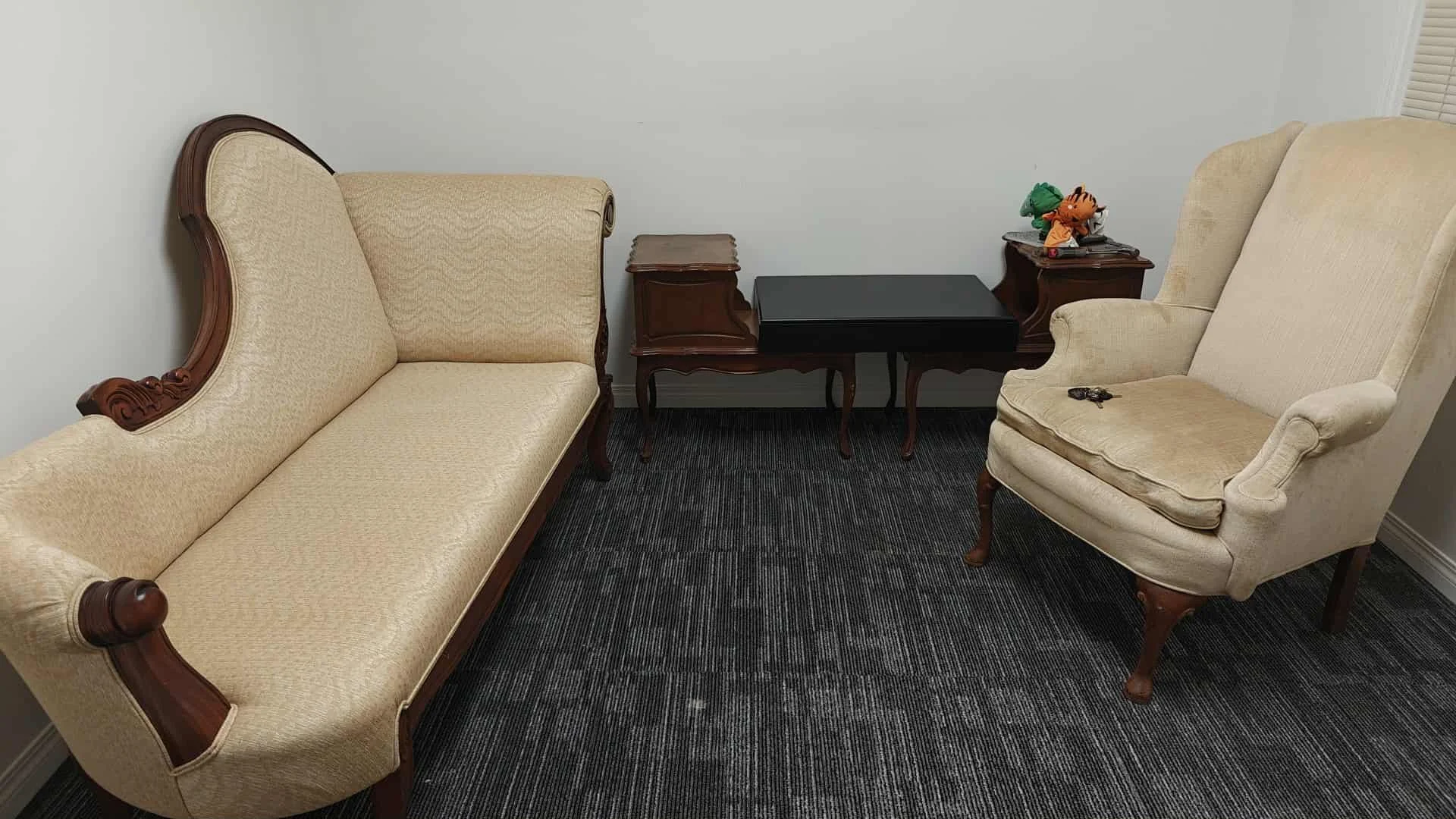 A vintage-style living room setup with a beige sofa, a beige armchair, dark wooden side tables, and a black coffee table on a dark carpet with horizontal stripes. The side tables hold some items, including plush toys and miscellaneous objects.