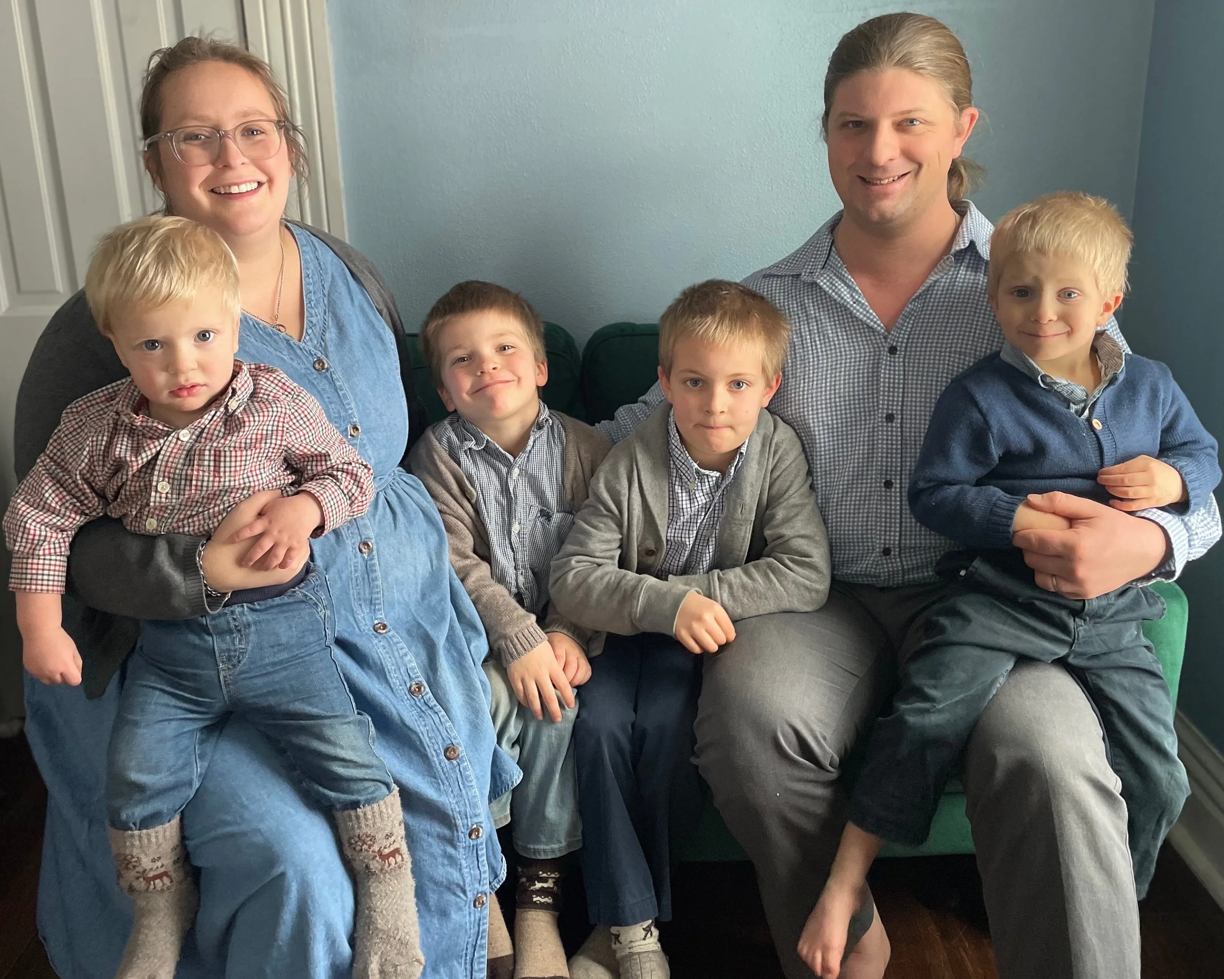 A family portrait of six members sitting on a sofa in a living room. Two adults, one woman and one man, are sitting in the middle, surrounded by four young boys. The woman is wearing glasses and a blue dress, and the man has long hair tied back, wearing a checkered shirt. The boys are dressed in collared shirts and cardigans, and one boy is being held by a woman on her lap. The background features a blue wall and a white door.