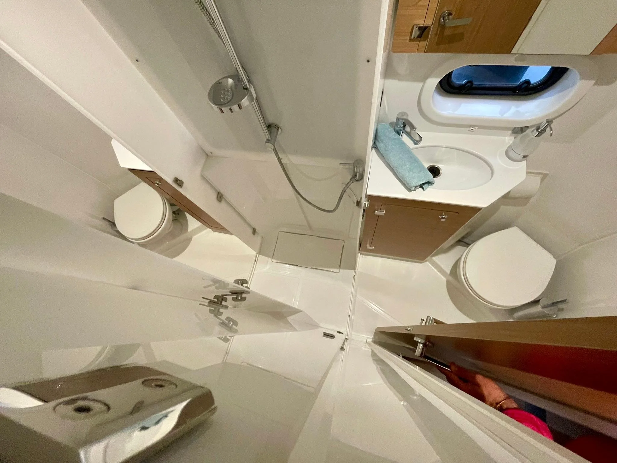 Small bathroom with a toilet, small sink with a blue towel, mirror, window, and a shower area with a handheld showerhead in a confined space.