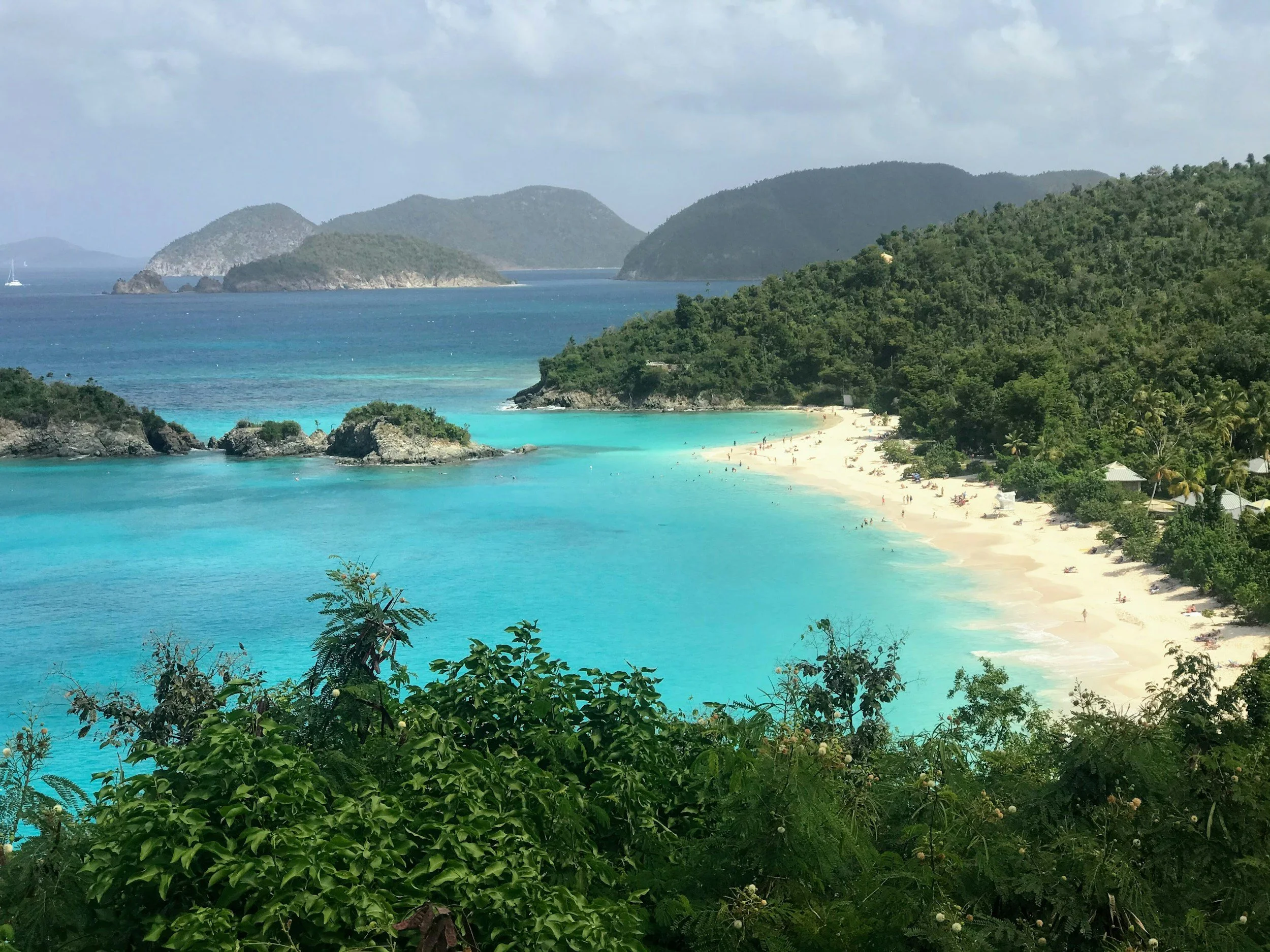 A tropical beach with white sand, turquoise waters, and lush green hills in the background, with small islands visible in the distance.