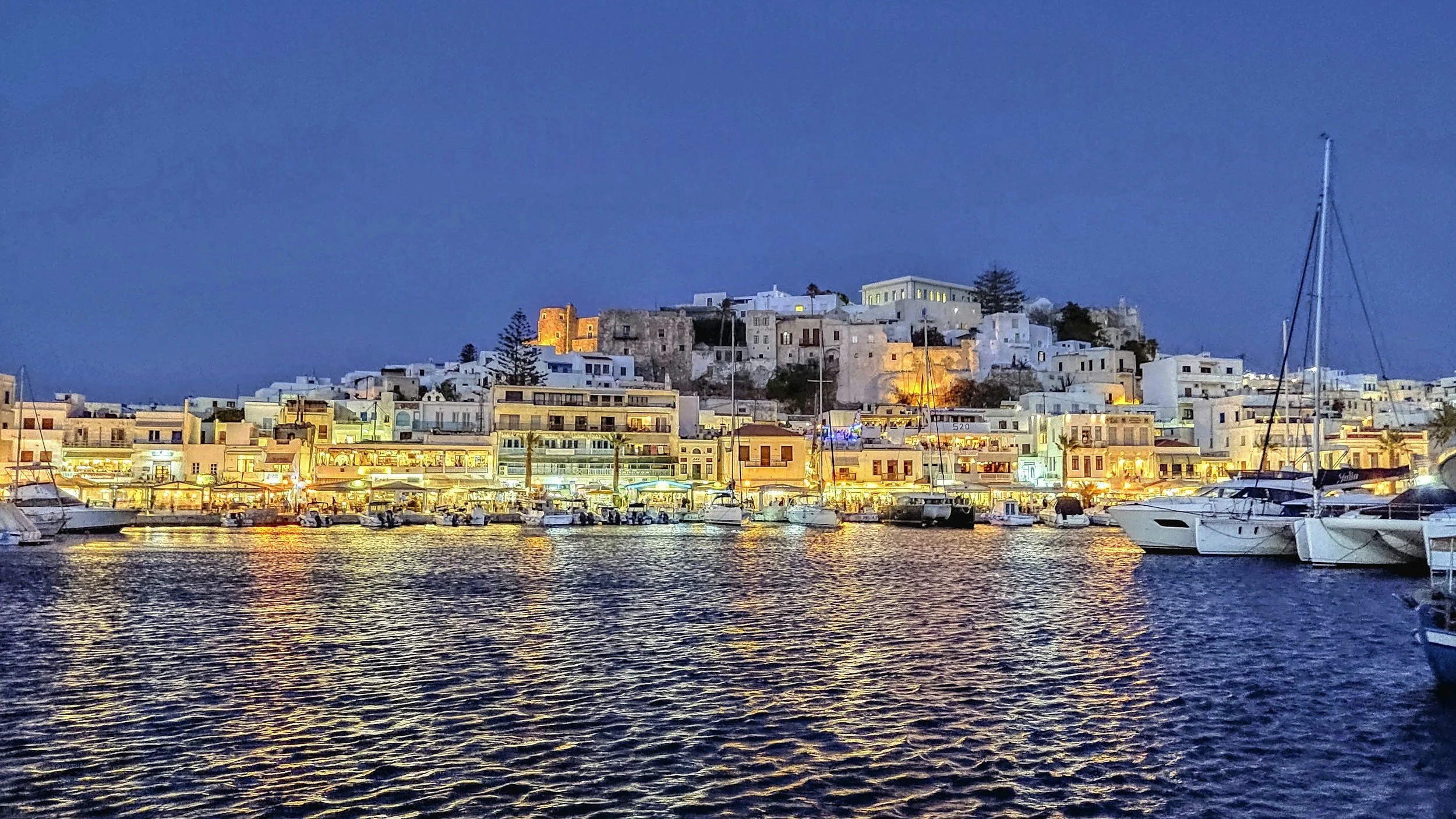 Night view of a harbor with boats and yachts docked, illuminated buildings along the waterfront, and a hillside town with white buildings and some lit structures, under a deep blue sky.