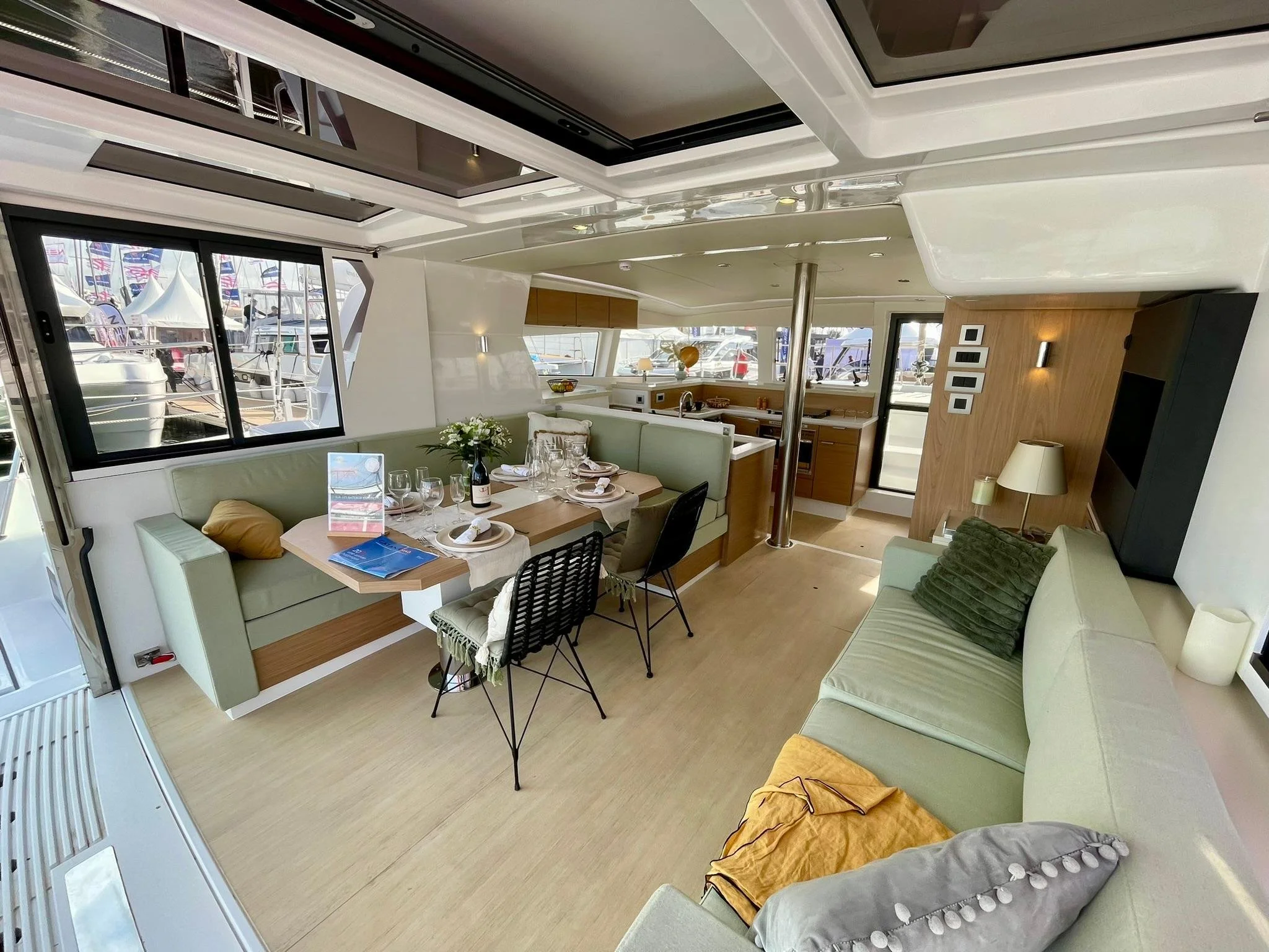 Interior of a yacht with a dining area set with plates, glasses, and a small flower arrangement, surrounded by light green seating, with large windows showing a marina with boats outside.