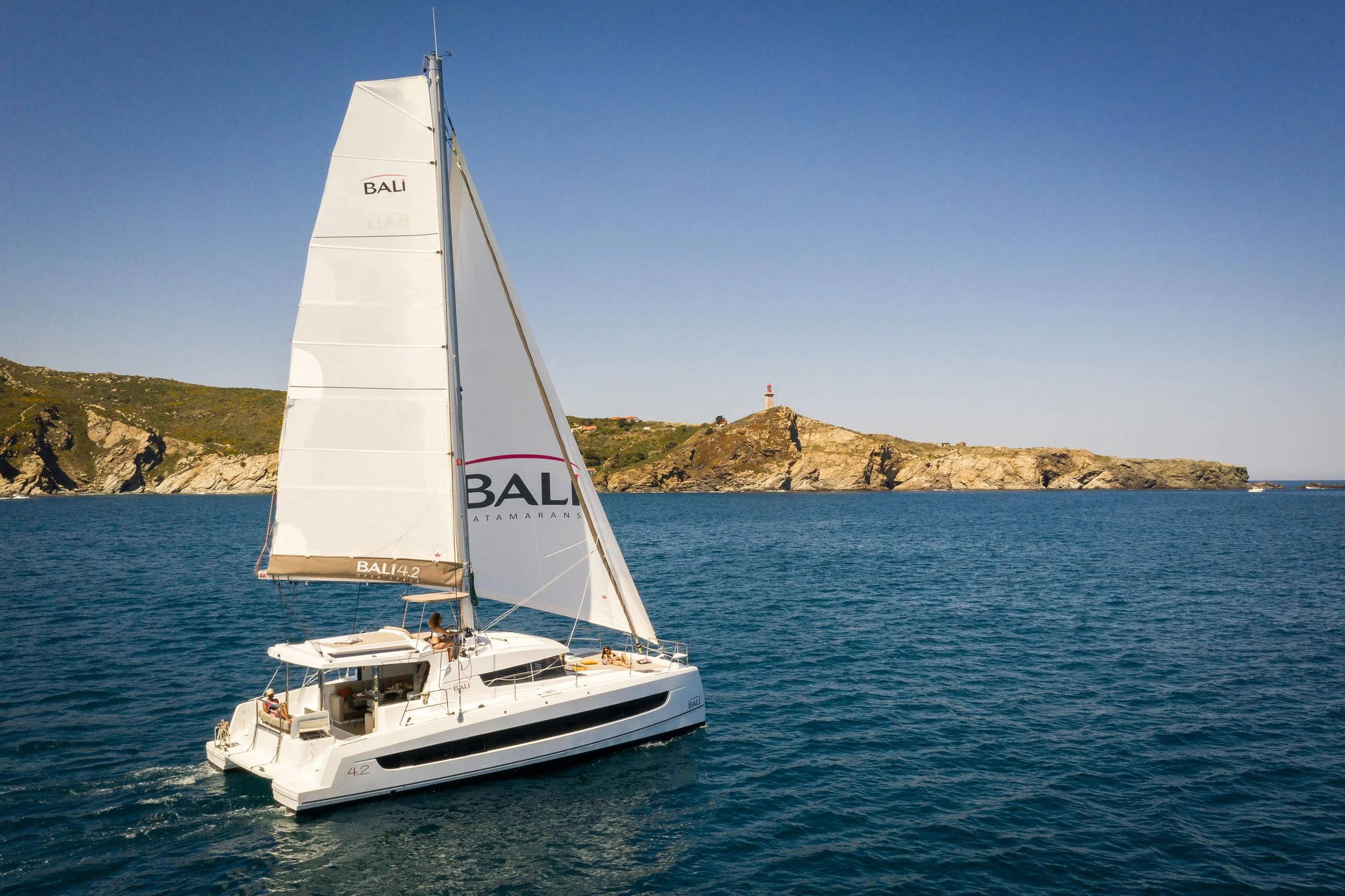 A sailboat with white sails on a calm blue sea, near a rocky island with a lighthouse on top, under a clear blue sky.