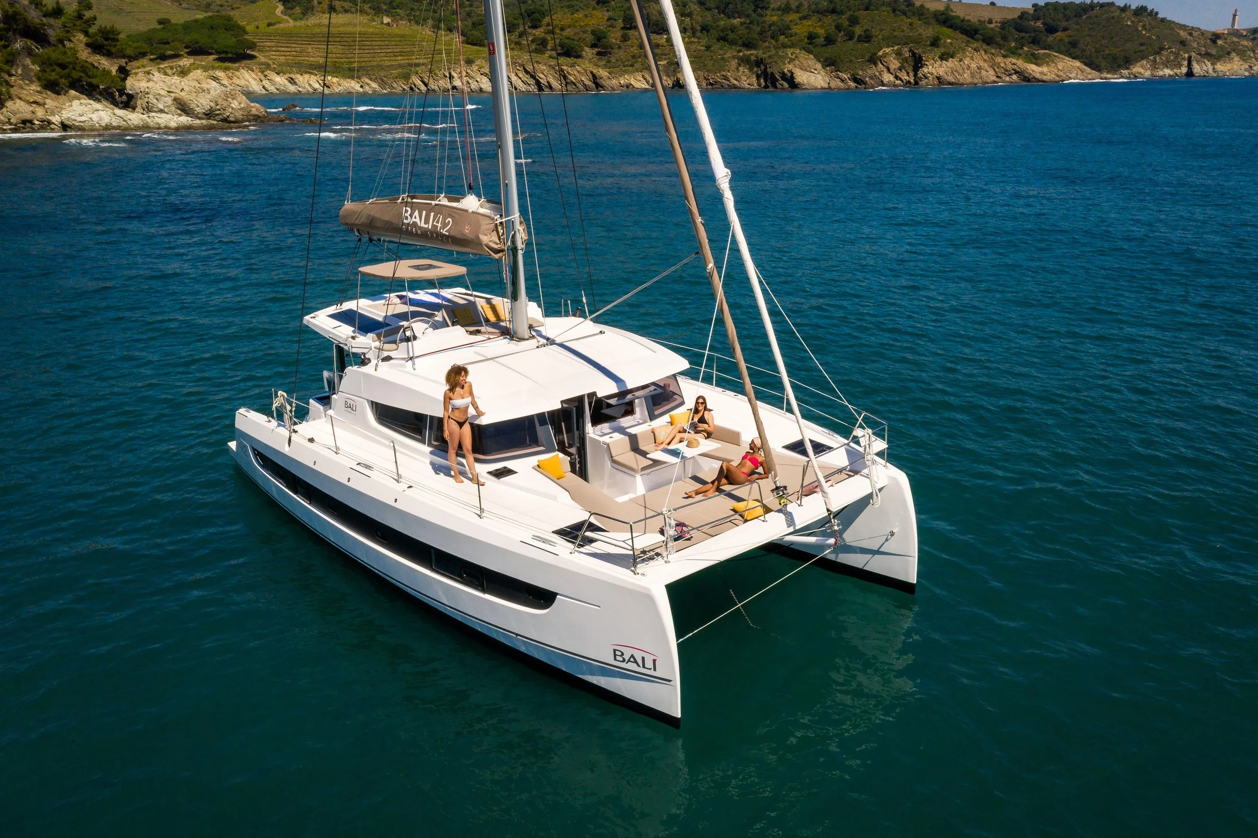 A white sailing yacht named BALI moored in clear blue water near a rocky shore with green hills, featuring three women relaxing and enjoying the sunny weather on deck.