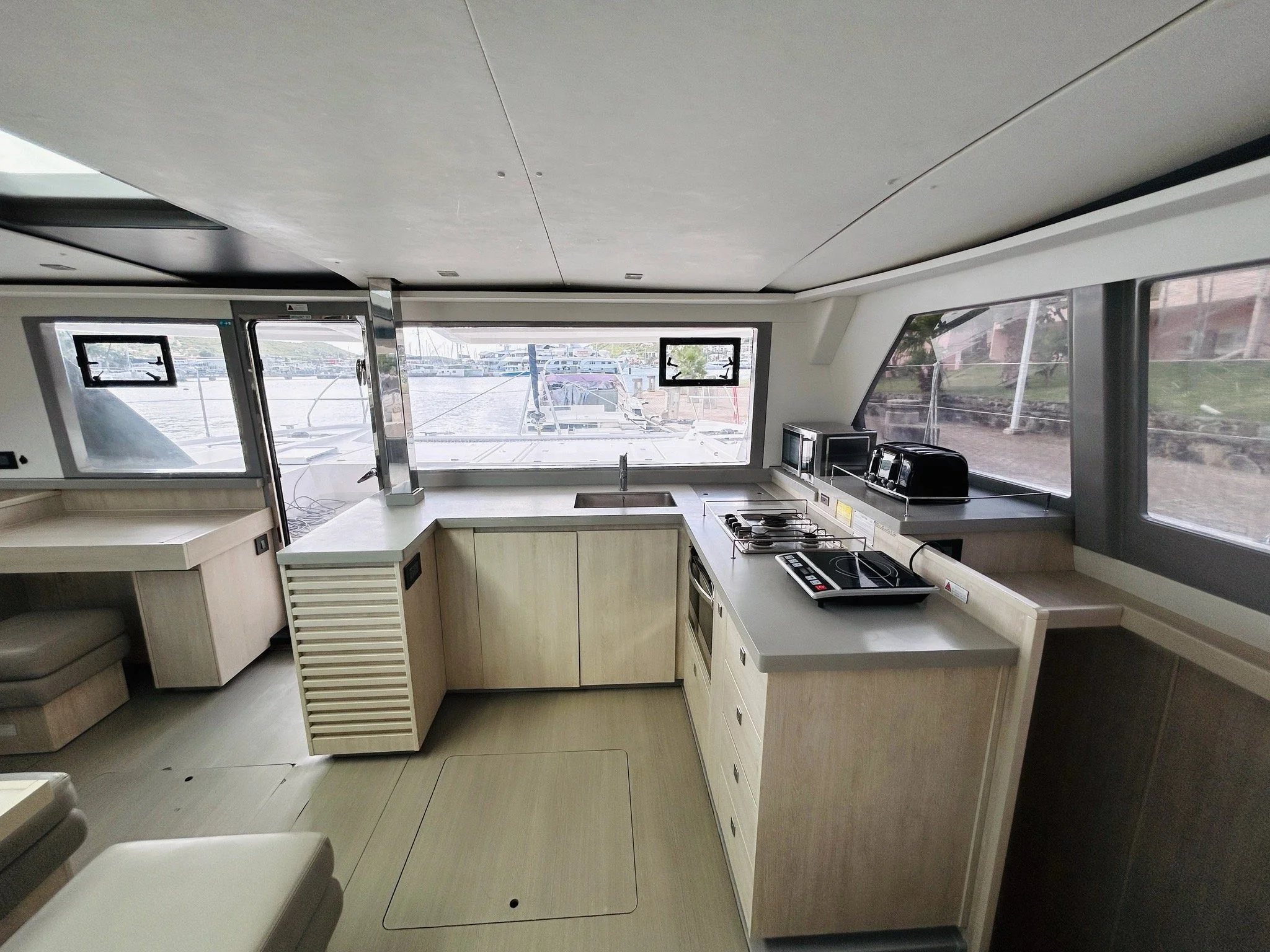 Interior of a modern boat's kitchen with light wood cabinets, a small sink, a stovetop, microwave, toaster, and large windows overlooking the marina.