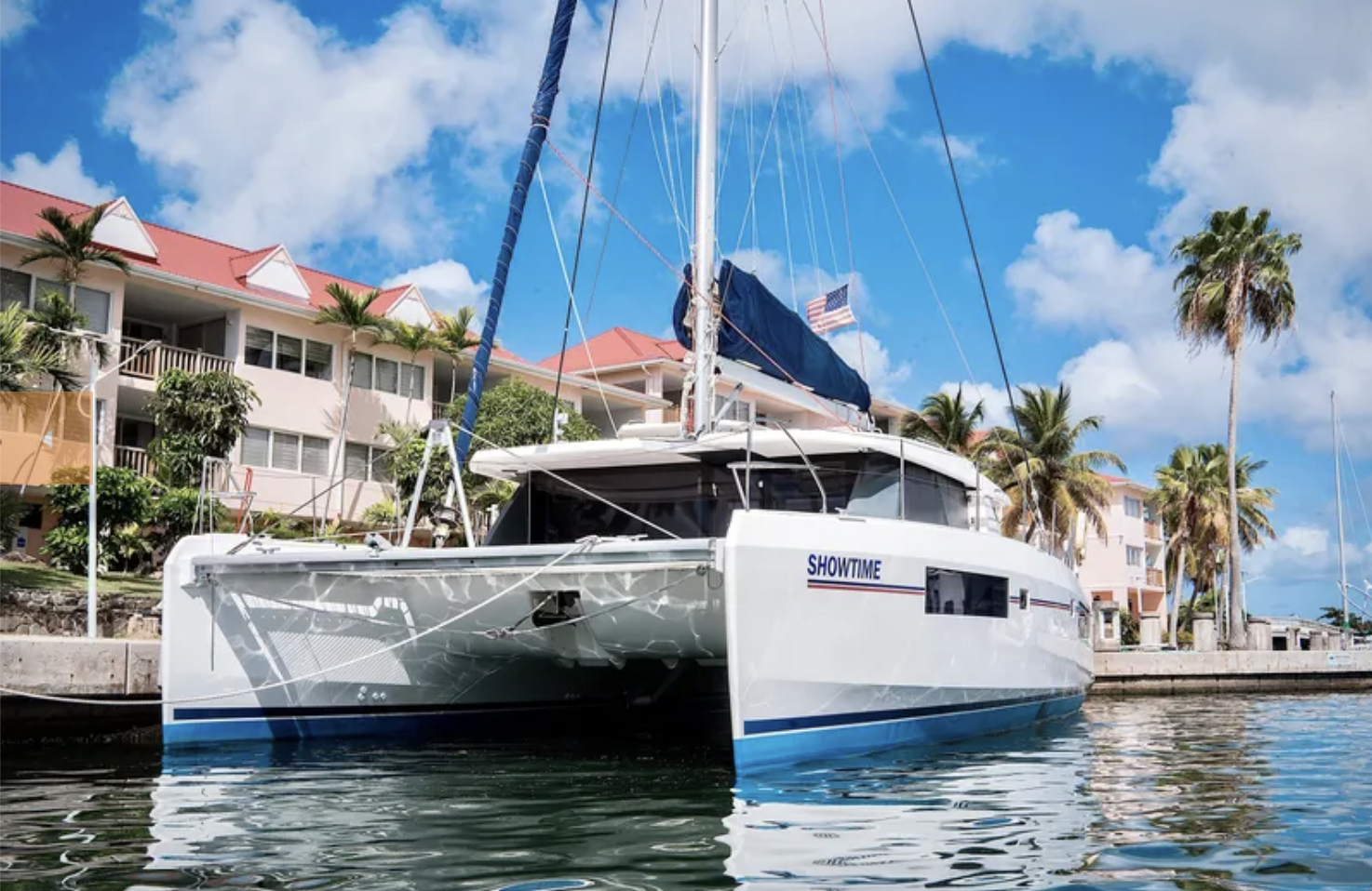 A sailboat named SHOWTIME docked at a marina near pink residential buildings and palm trees under a partly cloudy sky.