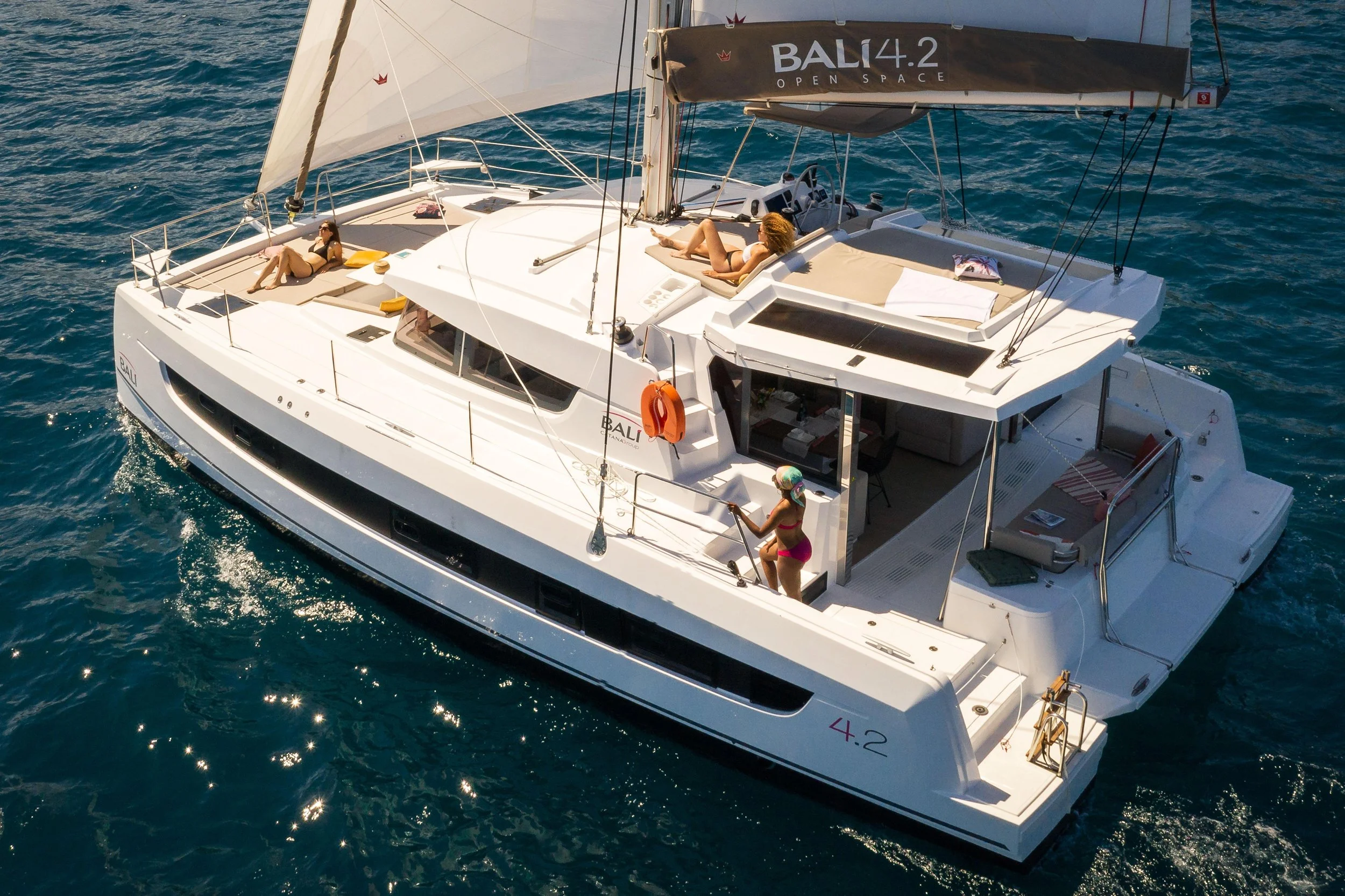A white luxury sailboat on the water with three women relaxing on deck, one standing near the entrance, and two lounging in sunbathing chairs on the upper deck.