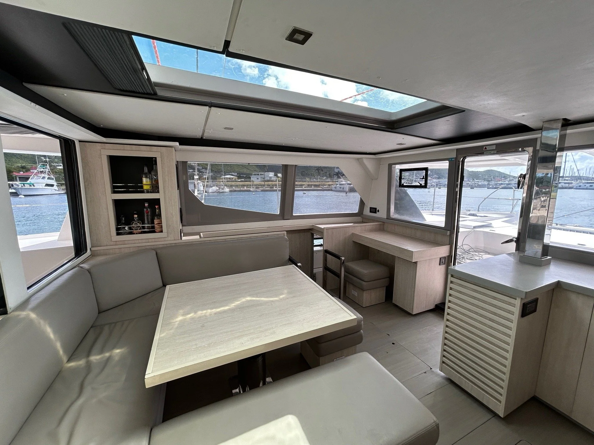Interior of a boat with a beige L-shaped seating area, a light wood table, a small bar cabinet with bottles, and large windows showing a marina with boats and water.