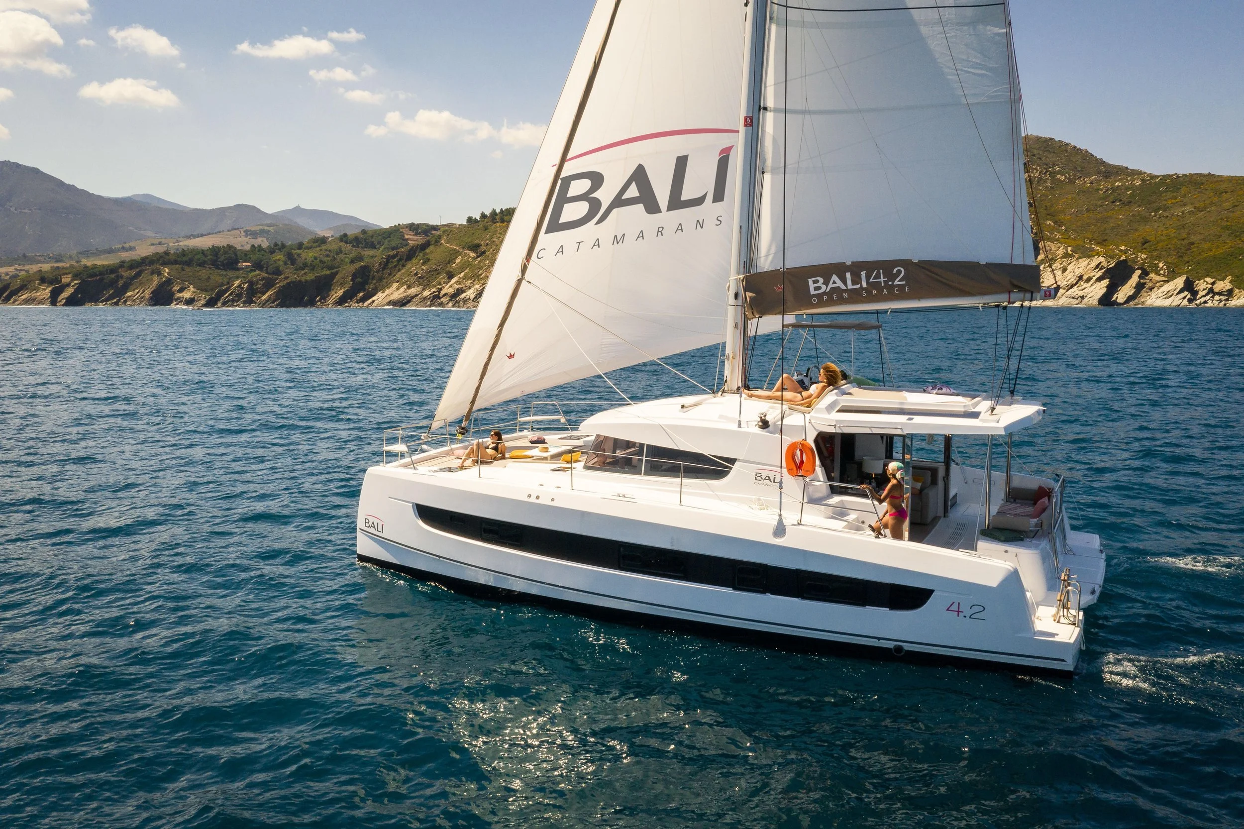 A white sailing yacht with people relaxing on deck, sailing on blue waters near rocky green land and mountains under a sunny sky.