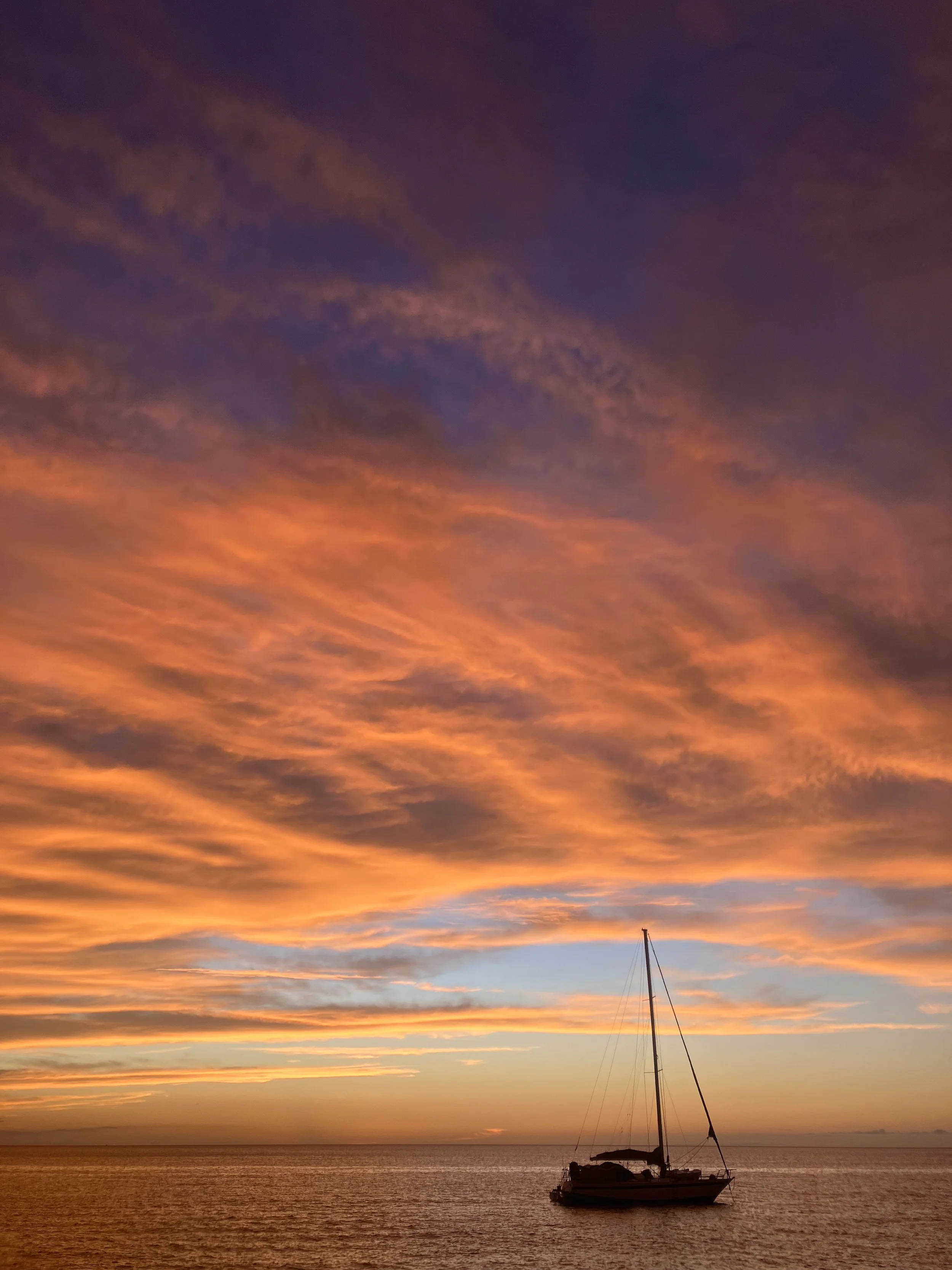 A sailboat floating on calm water during a colorful sunset with an orange, pink, and purple sky.