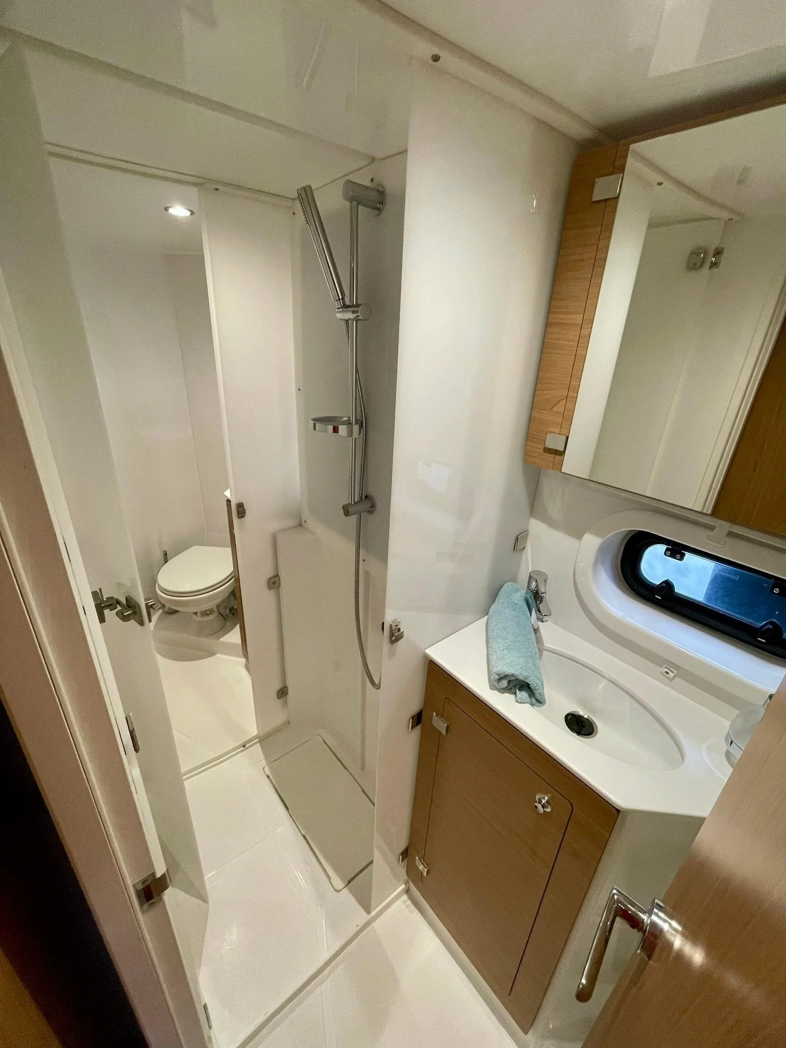 Small bathroom with shower area, toilet, and vanity with mirror. The shower has a sliding door, a handheld showerhead, and a small shelf. The vanity has a sink with a towel on it, a mirror above, and wooden cabinetry.