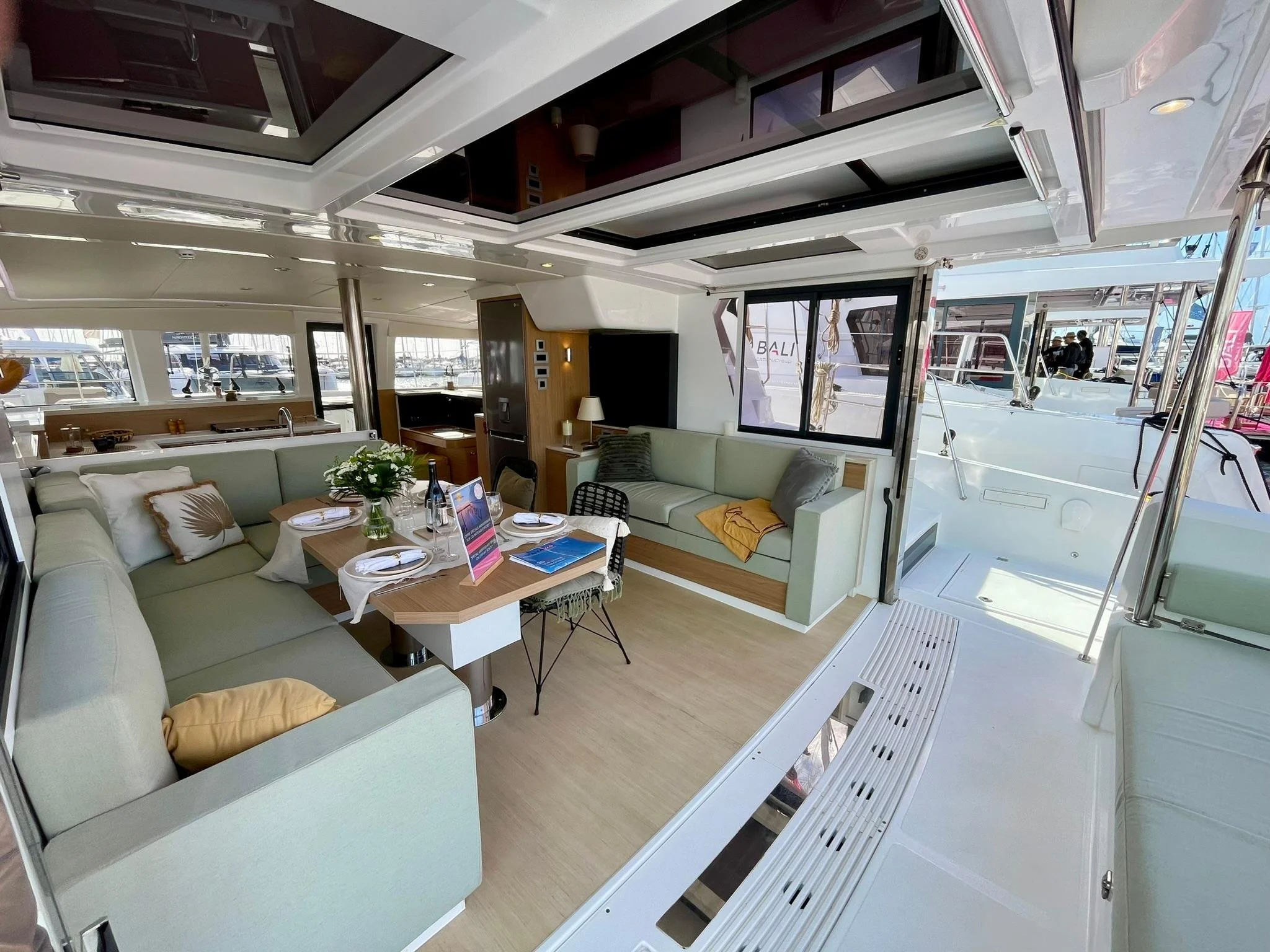 Interior of a luxury yacht with a dining area featuring a table set with plates, glasses, and a vase of flowers, surrounded by light green couches, with windows overlooking a marina full of boats.