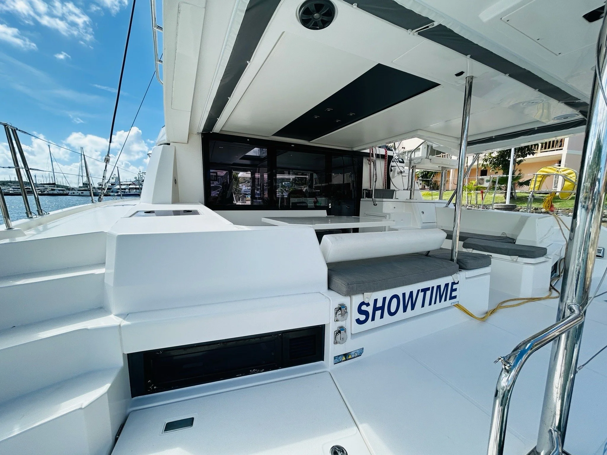 View of a luxury yacht's deck with seating and a sign that reads 'SHOWTIME'; there are cushions, stainless steel railings, and a marina with boats and a building in the background.
