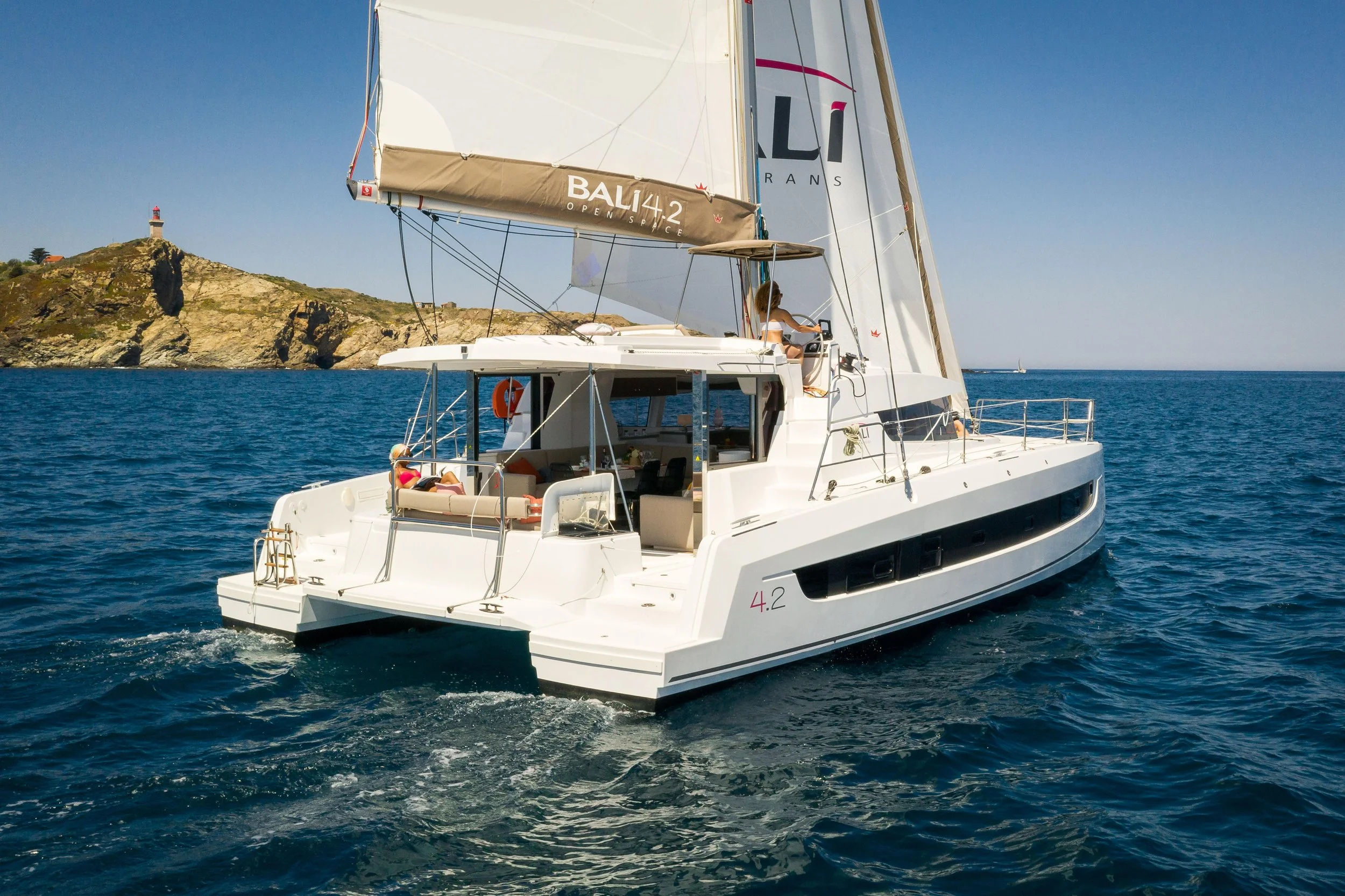 A white sailing yacht on the water near a rocky coastline with a lighthouse on a hill in the background.
