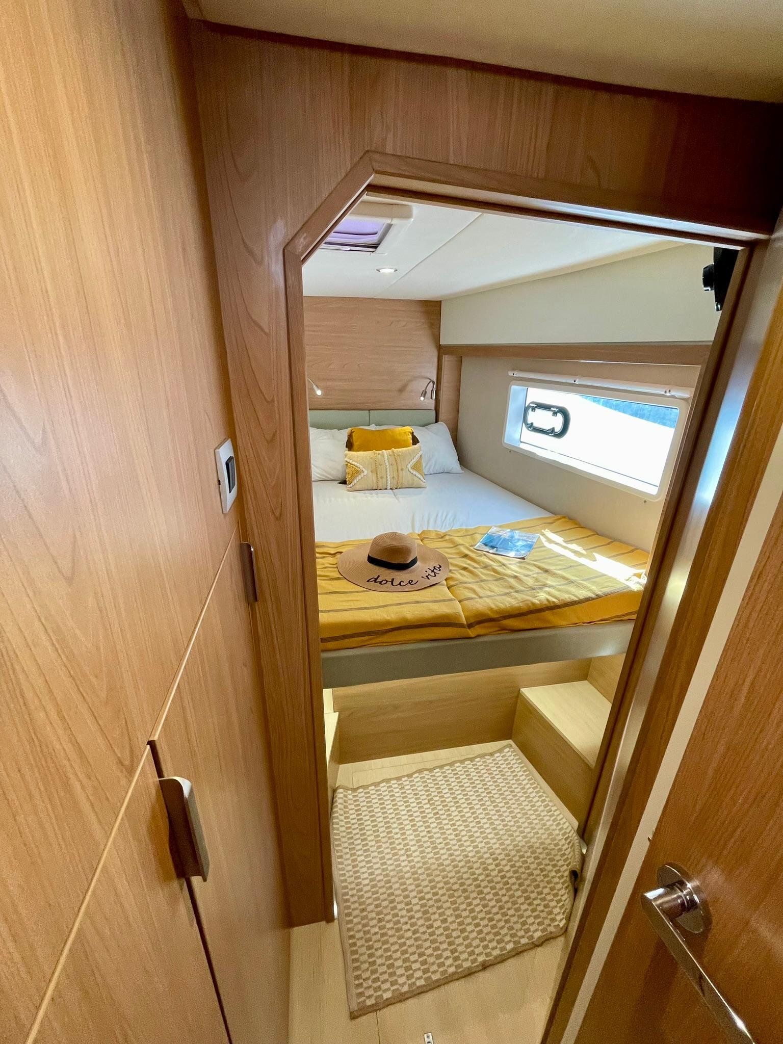 A cozy bedroom on a boat with a bed covered in yellow striped bedding, decorative pillows, a hat with 'dolce vita' embroidered, and a magazine. There is a window on the right and a small ledge or step at the foot of the bed. The room has wood panelin