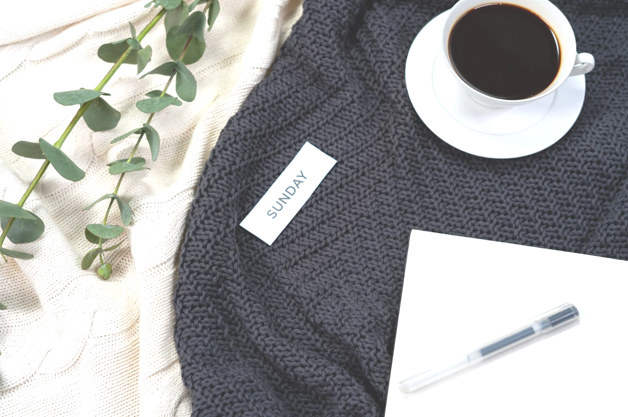 A cup of black coffee on a saucer, a white blank notebook, a clear pen, a gray knitted blanket, a white tag labeled "Sunday," a cream-colored textured cloth, and a green leafy branch.
