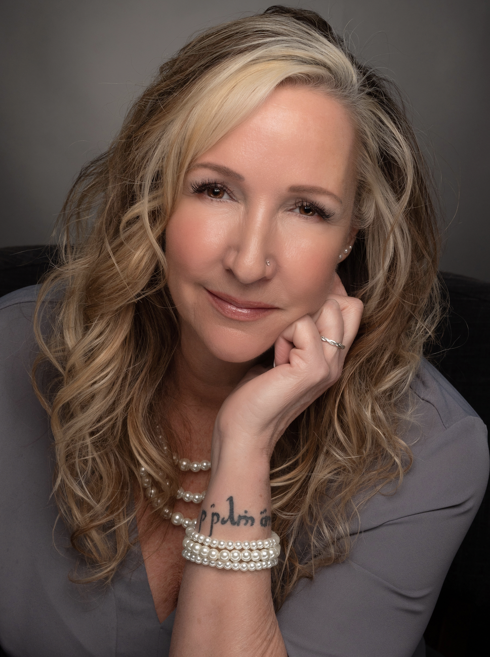 A woman with wavy blonde hair, wearing a grey top, pearl necklace and bracelet, with a nose ring, posing with her right hand resting on her chin, smiling softly at the camera.