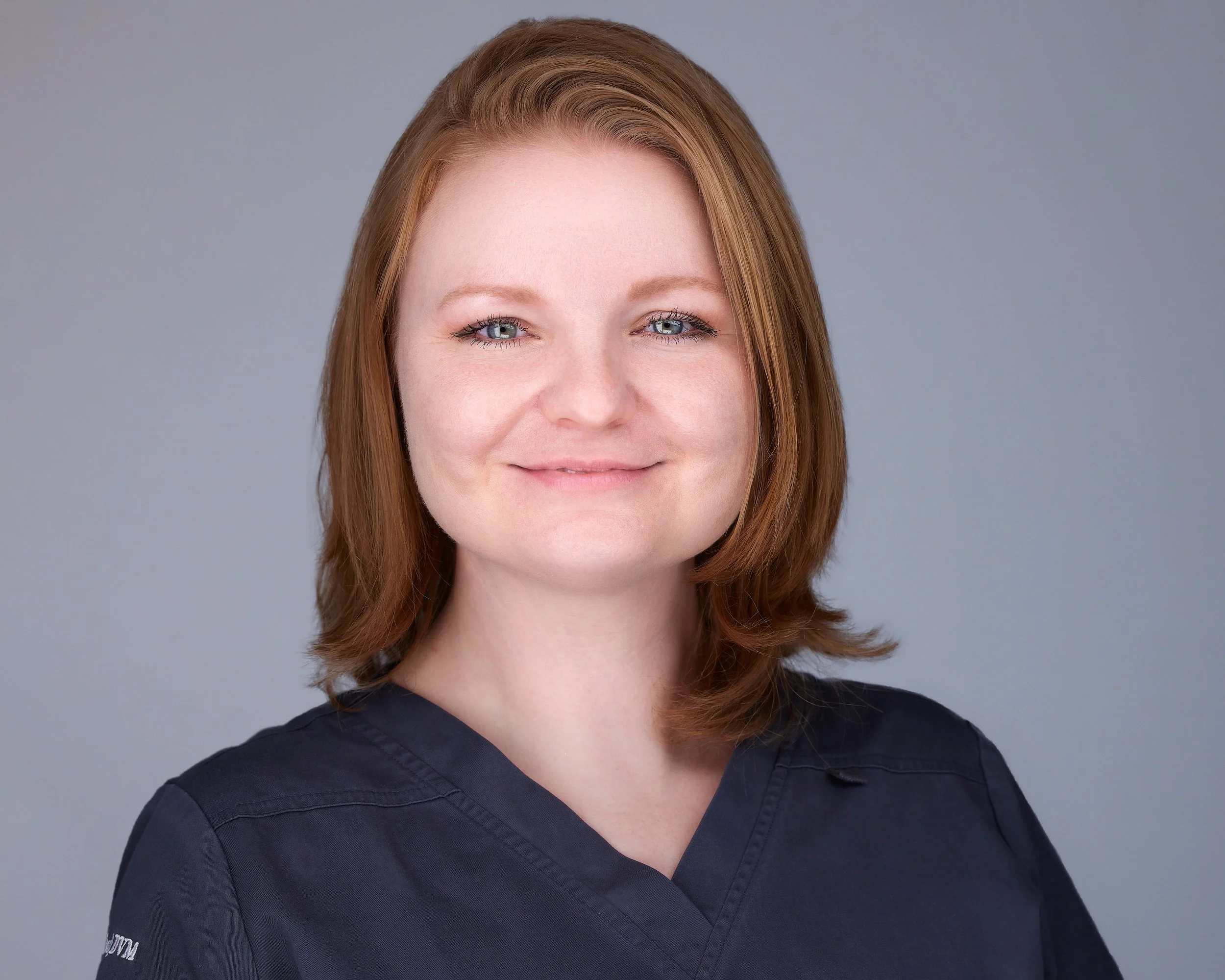A woman with shoulder-length red hair, wearing a dark navy medical scrubs, smiling at the camera against a plain grey background.