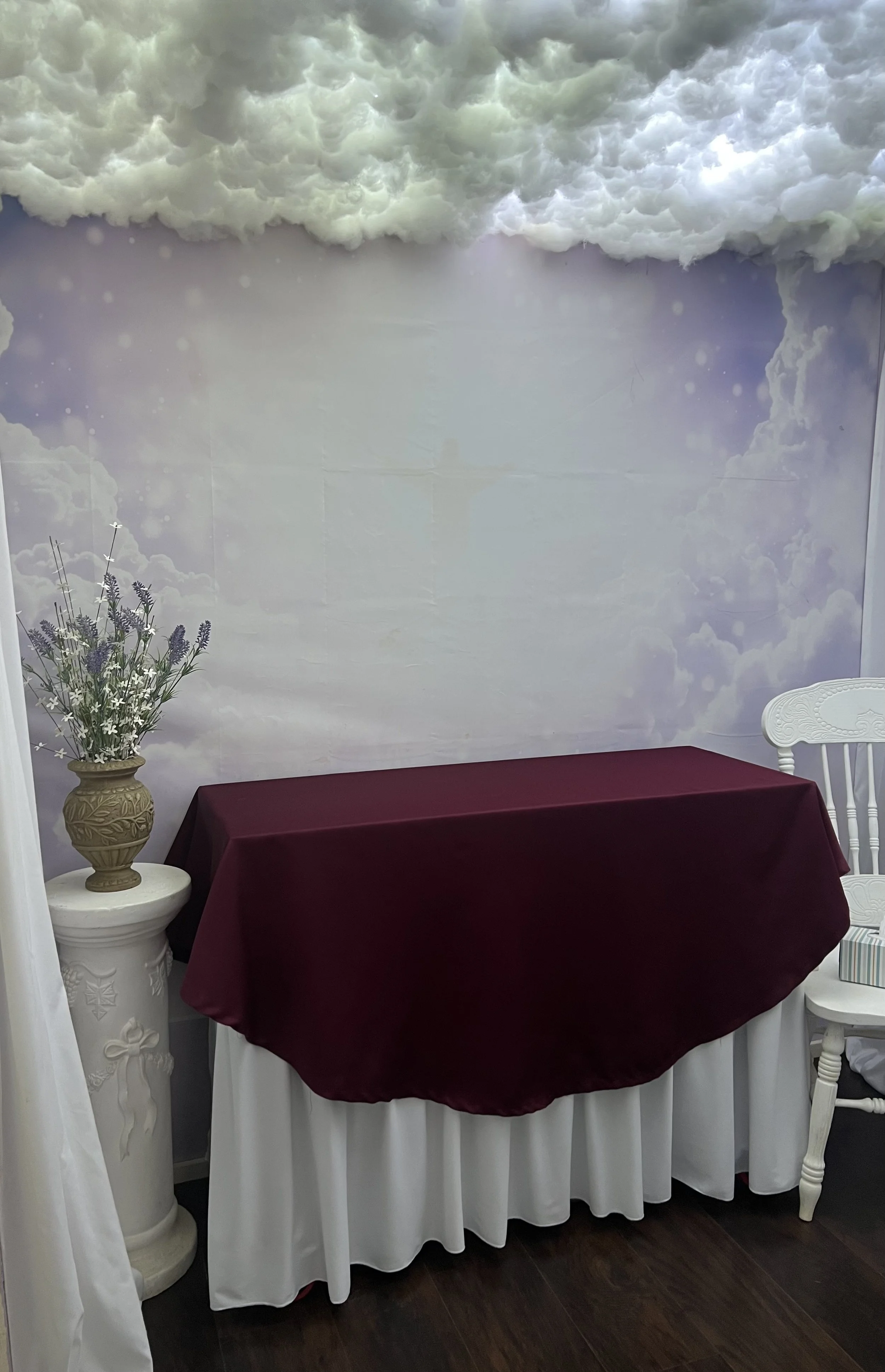 Indoor scene with a decorated wall featuring clouds and sky, a table with a burgundy tablecloth, a white floral-patterned table skirting, a white chair on the right, and a tall vase with lavender and white flowers on a white pedestal on the left.