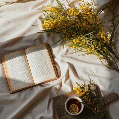 Open book, yellow flowers, and a cup of tea on a cloth-covered surface.