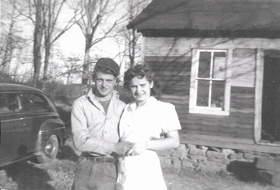 A black and white photo of a young man and woman standing outdoors, smiling, with a vintage car parked nearby and a wooden house in the background.