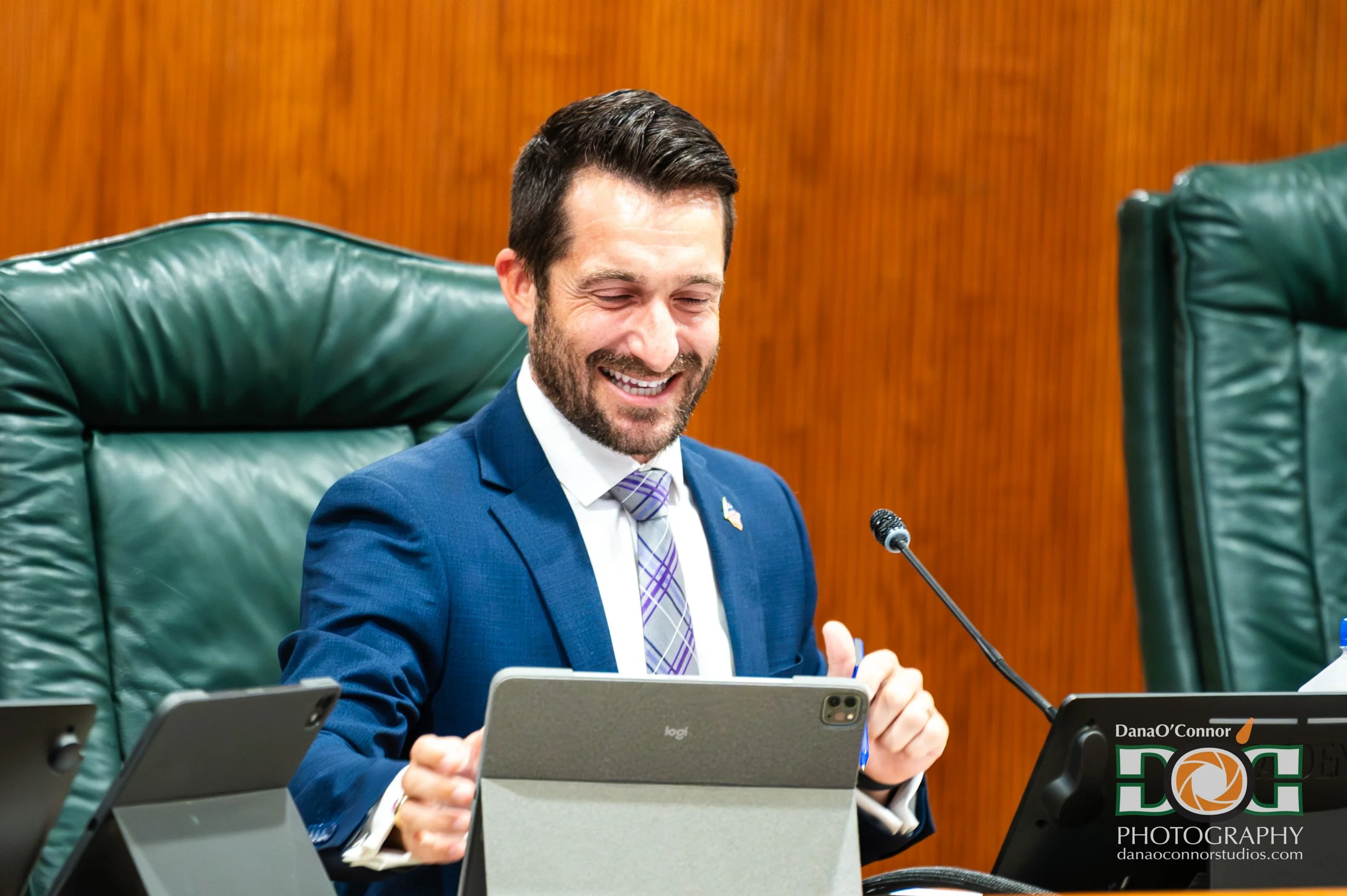 A man in a blue suit and purple tie smiling and speaking at a table with multiple electronic devices in front of him, in a conference room with wooden walls.