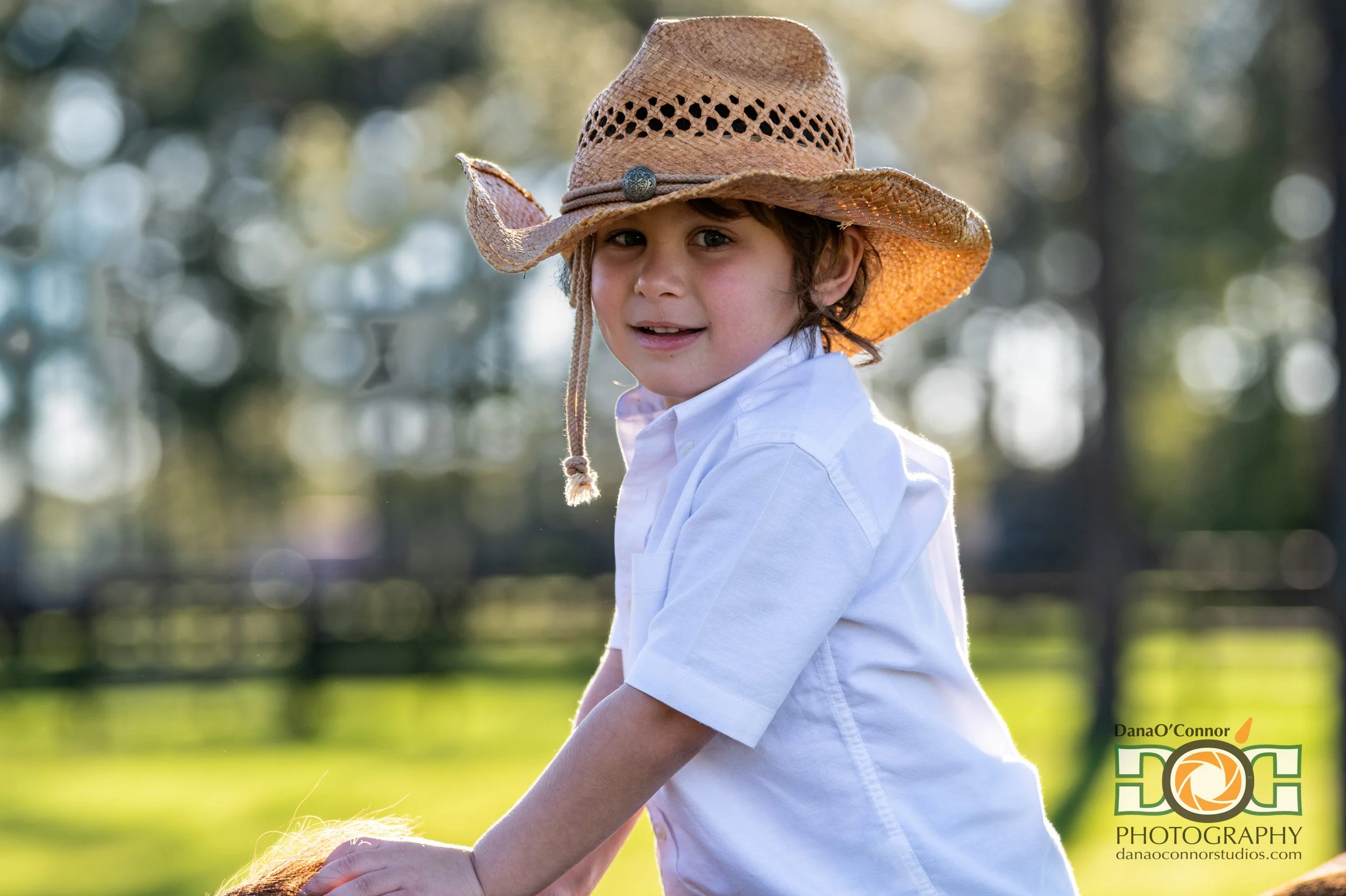 A young boy with curly hair wearing a straw cowboy hat and a white collared shirt, outdoors in a sunny park with blurred trees in the background.