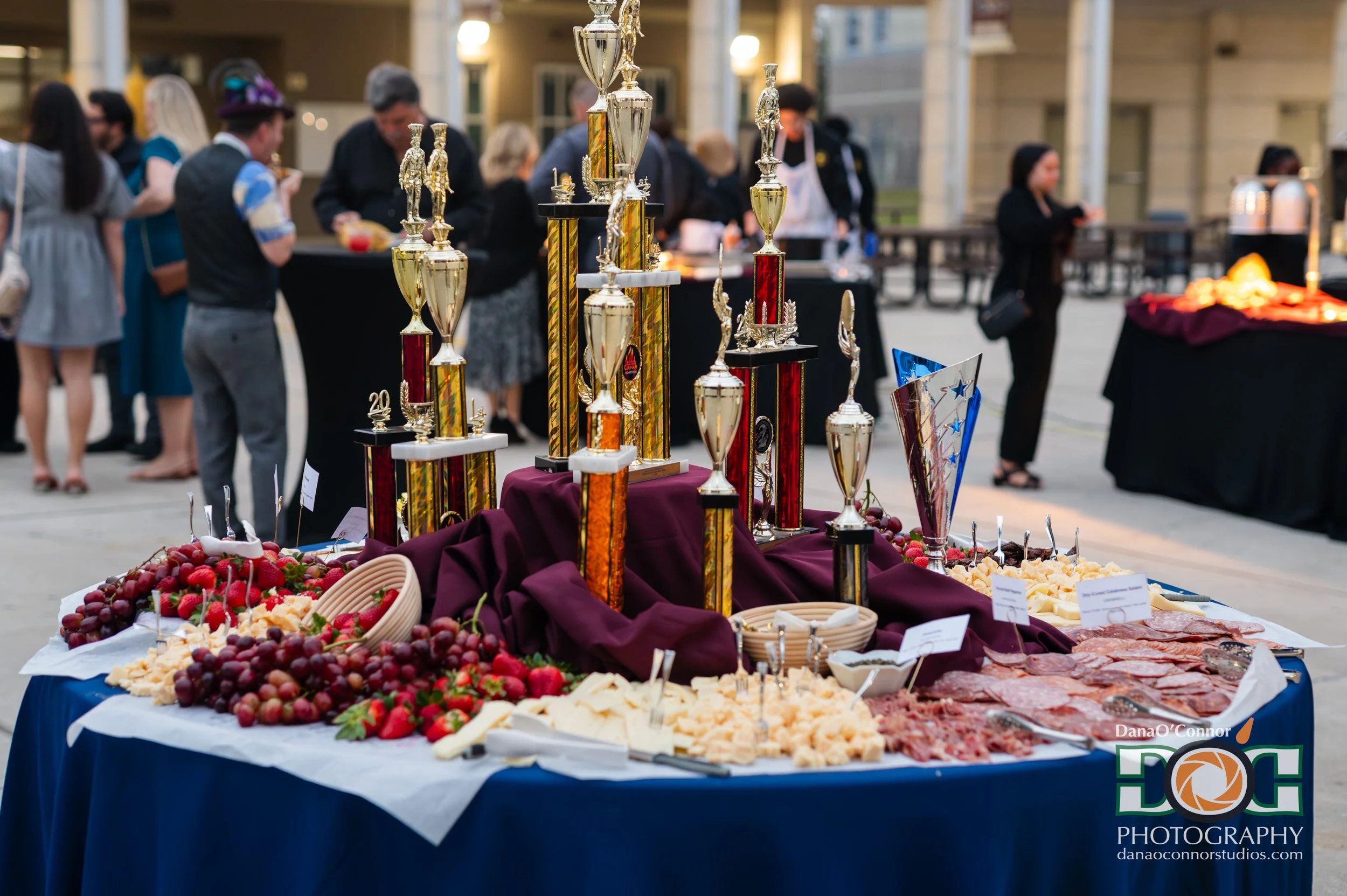 A buffet table with various cheeses, strawberries, grapes, and meats, decorated with trophies displayed behind the table at an outdoor event.