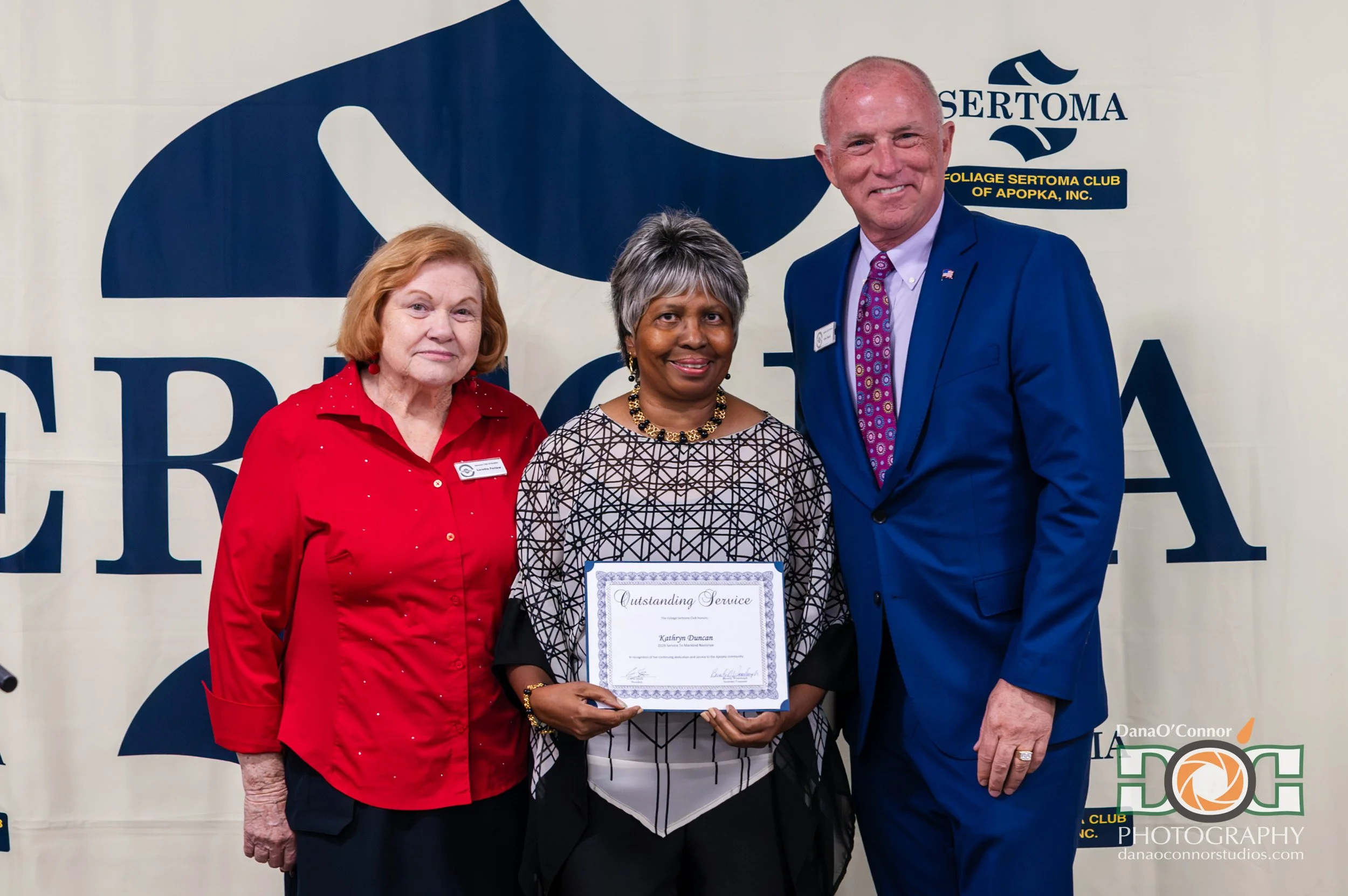 Three people standing together, with one woman holding an award certificate labeled 'Outstanding Service.' The woman in the center has short gray hair and is wearing a patterned black and white top. The woman on the left has red hair, wearing a red s