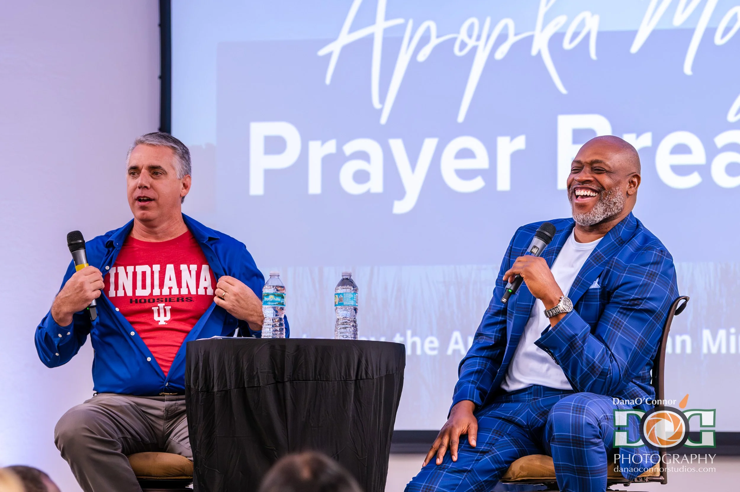 Two men speaking at an event, one wearing an Indiana University shirt and the other in a blue plaid suit, sitting at a table with water bottles against a background screen that reads 'Prayer' and other words.