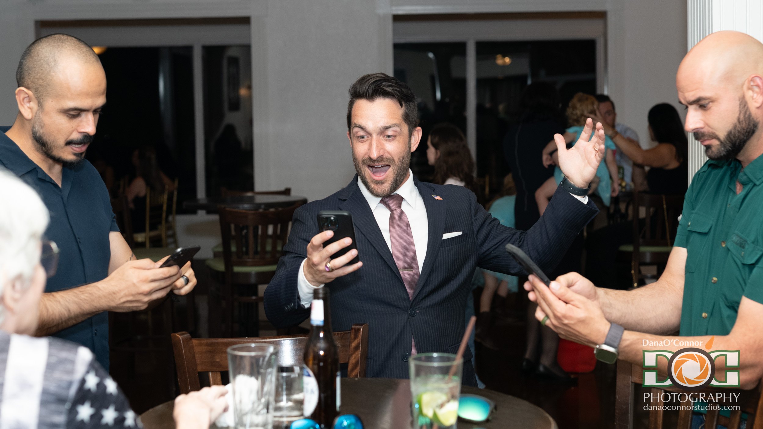 A man in a suit and tie at a social gathering, smiling and gesturing excitedly while holding a smartphone, surrounded by other people using their phones and chatting in a restaurant or bar setting.