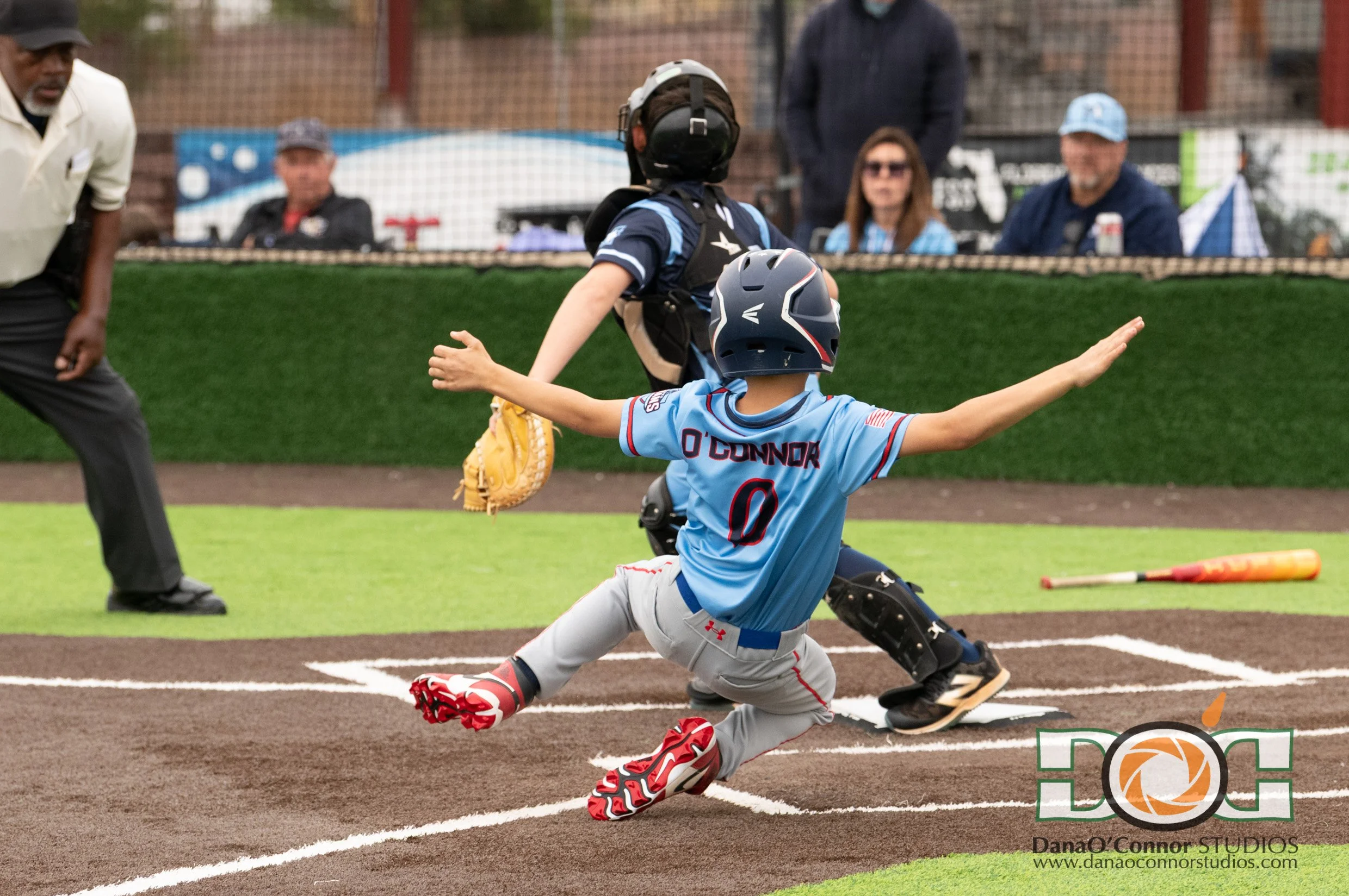 A baseball player in a blue uniform slides into home plate while the catcher in gray and black gear tries to tag him out. An umpire and other spectators are in the background.