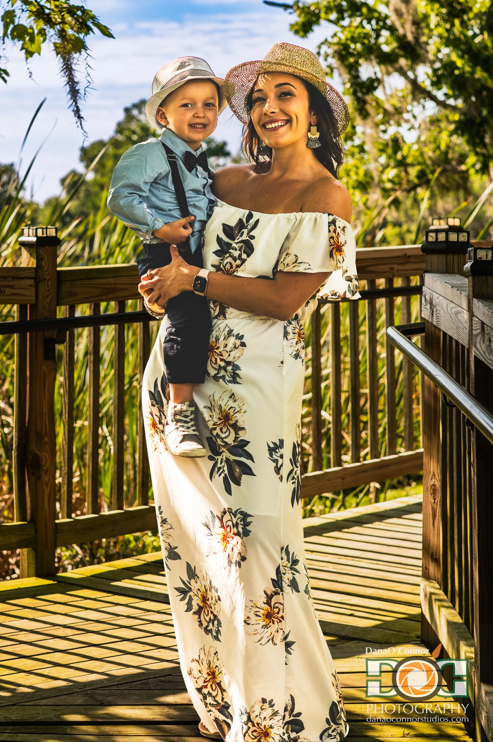 A woman and a young boy smiling outdoors on a wooden deck with lush greenery and trees in the background, wearing summer hats and floral attire.