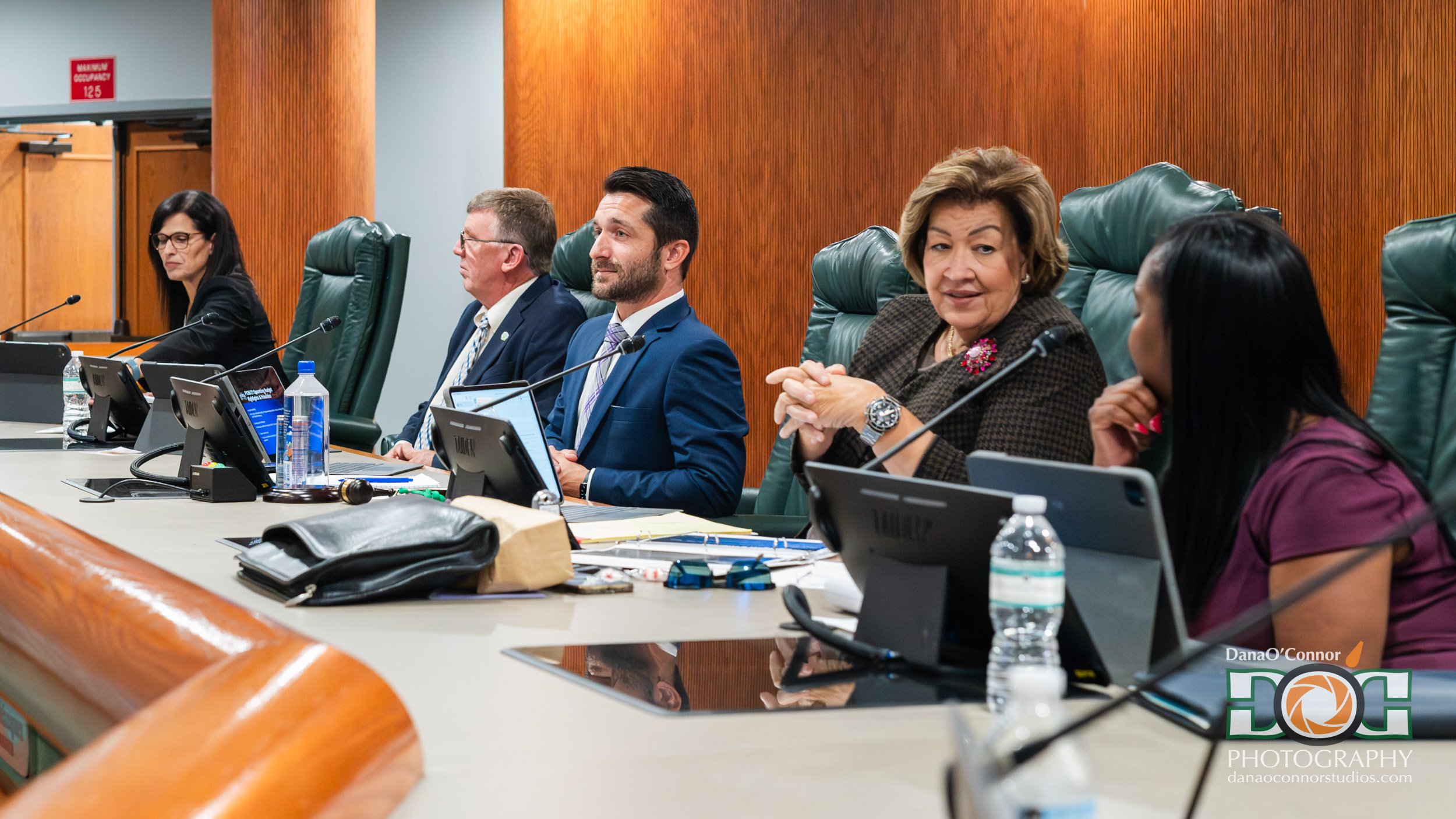 A group of diverse officials and professionals seated at a long conference table during a formal meeting, engaged in discussion, with microphones, laptops, water bottles, and documents in front of them.