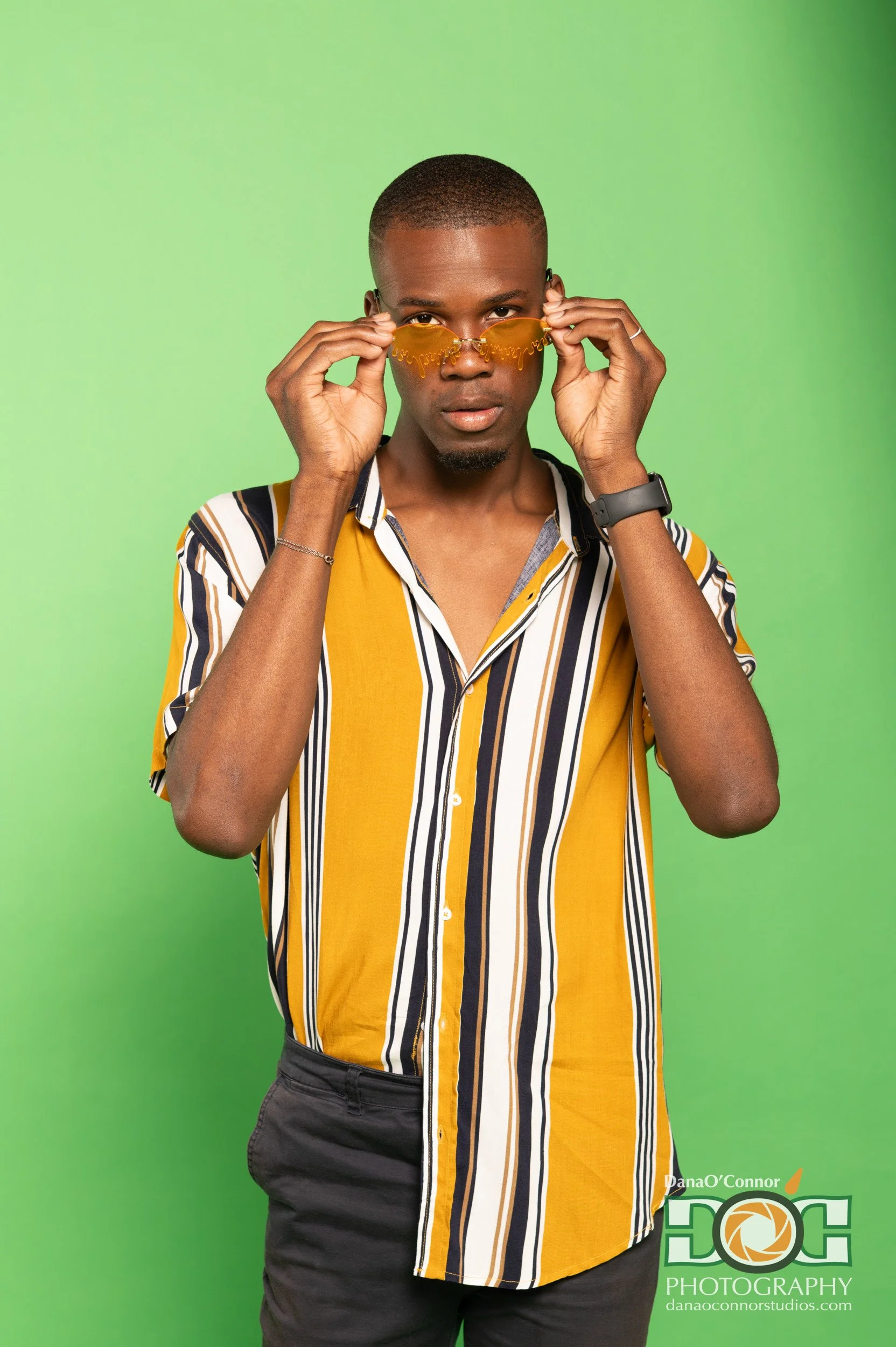 A young man with short hair and dark skin, wearing a yellow, black, and white striped short-sleeve shirt, black pants, and a gray wristwatch, holds yellow sunglasses with writing on the lenses with both hands, standing against a green background.