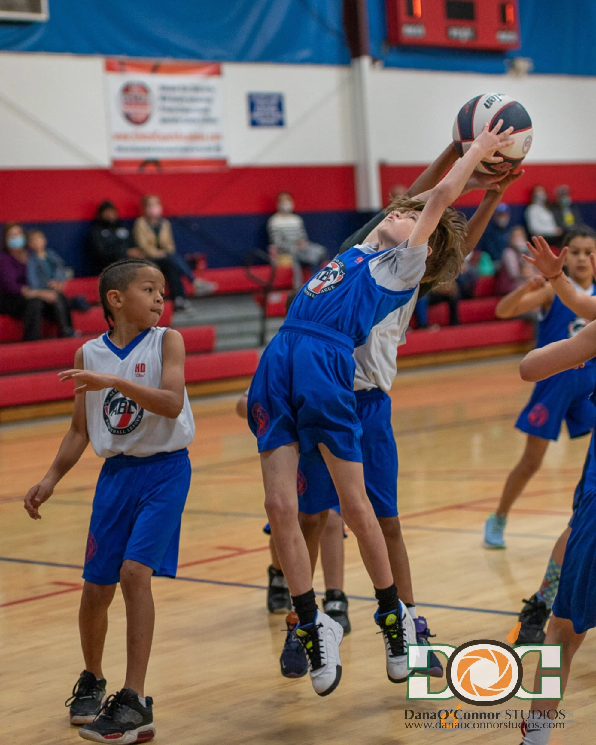 Children playing basketball in an indoor gymnasium, with a boy jumping to shoot or catch a basketball, others watching, and spectators sitting on bleachers in the background.