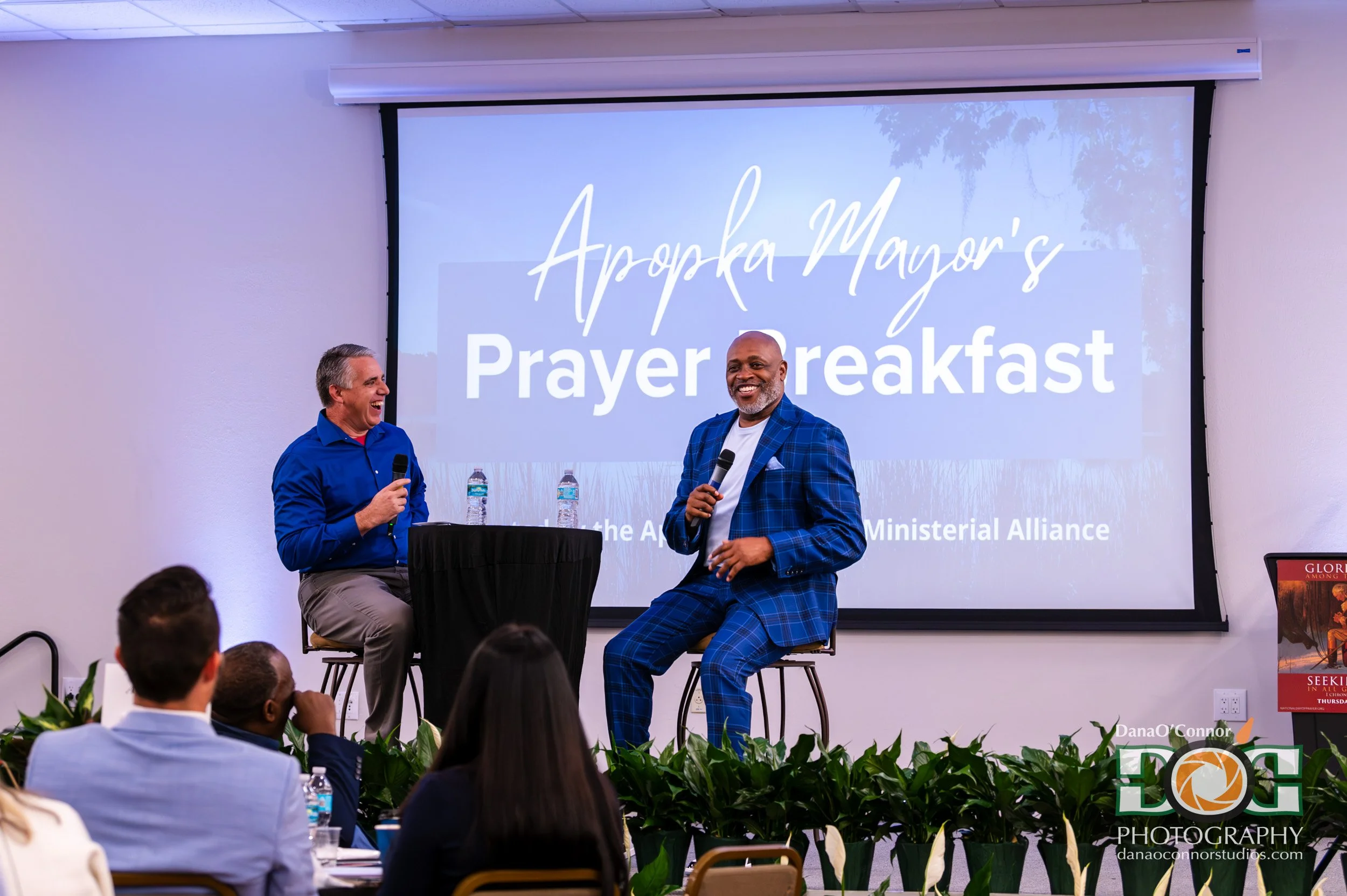 Two men on stage, one sitting and one standing, engaging in a discussion during a prayer breakfast event, with a large screen behind them displaying the event title, 'Apropos Mayor's Prayer Breakfast.' Audience members are seated in the foreground.