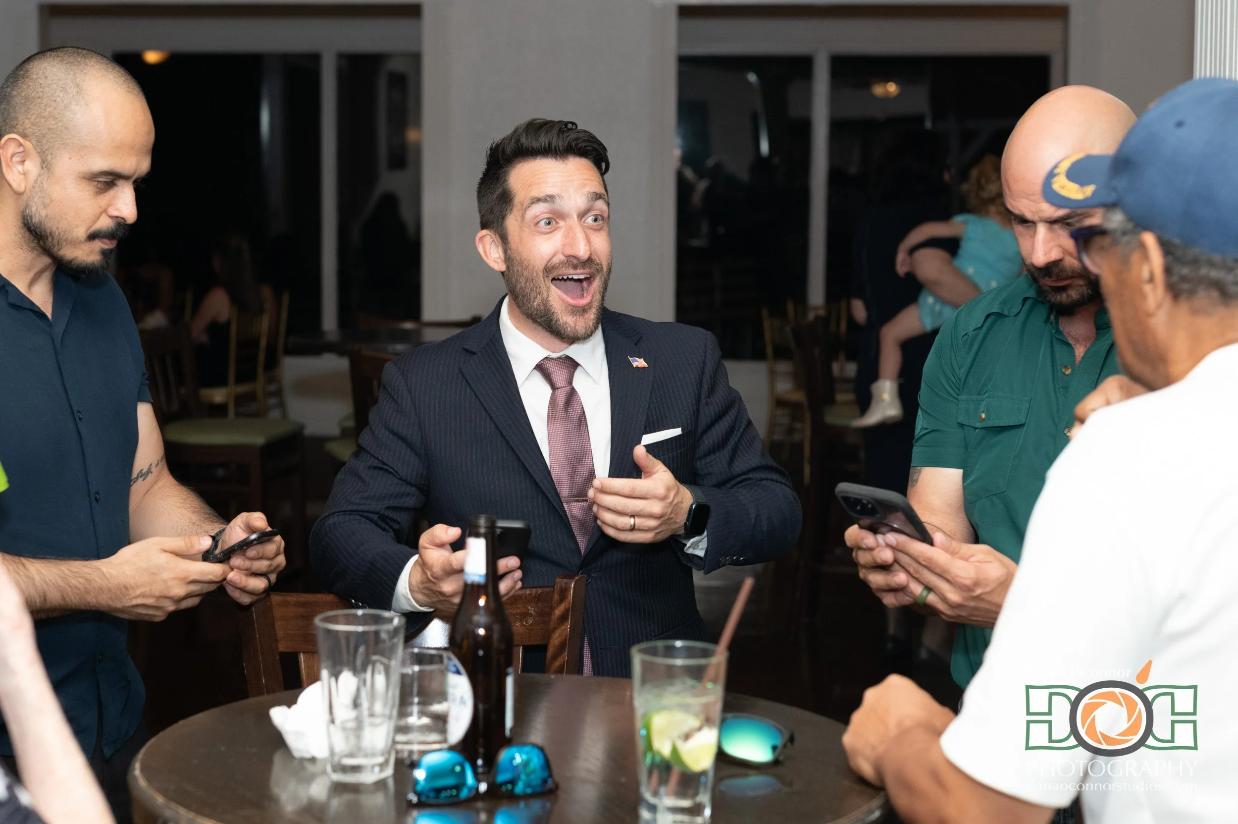 A group of four men gathered around a table, two of them are looking at smartphones, one is wearing a suit, and the others are dressed casually. They are in a restaurant or bar setting, with drinks and sunglasses on the table.