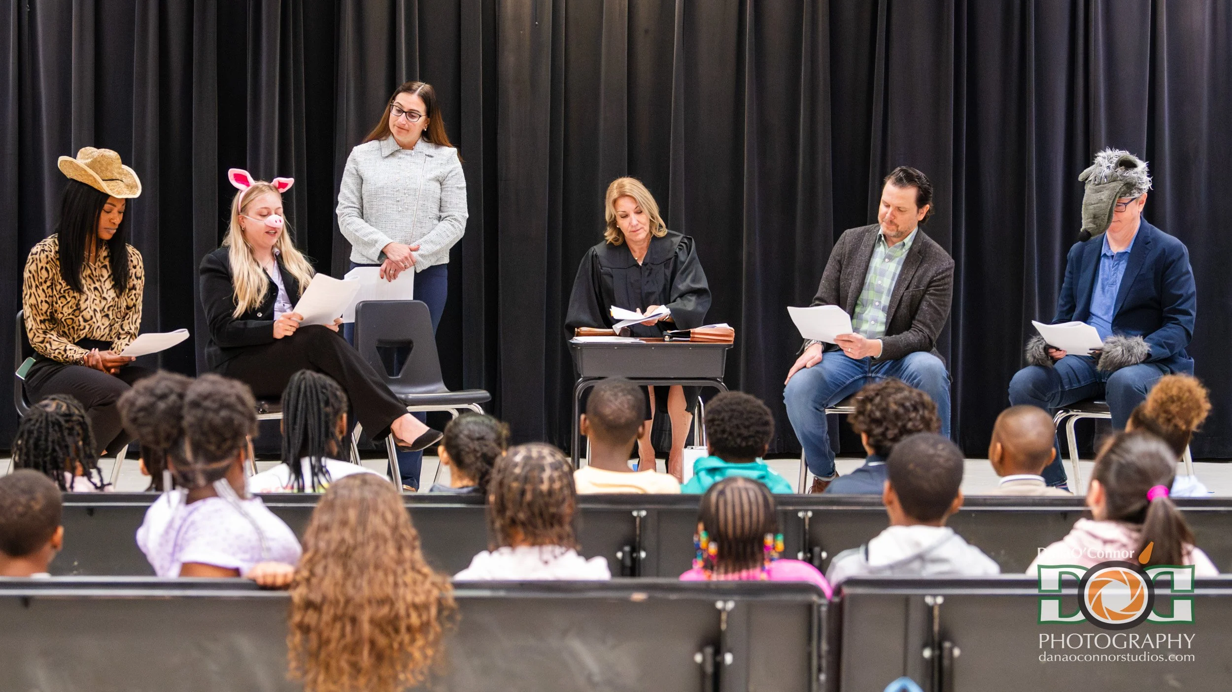 roup of six people on a stage reading from papers, some dressed in costumes, facing an audience of children with braids and pigtails, black curtain backdrop.