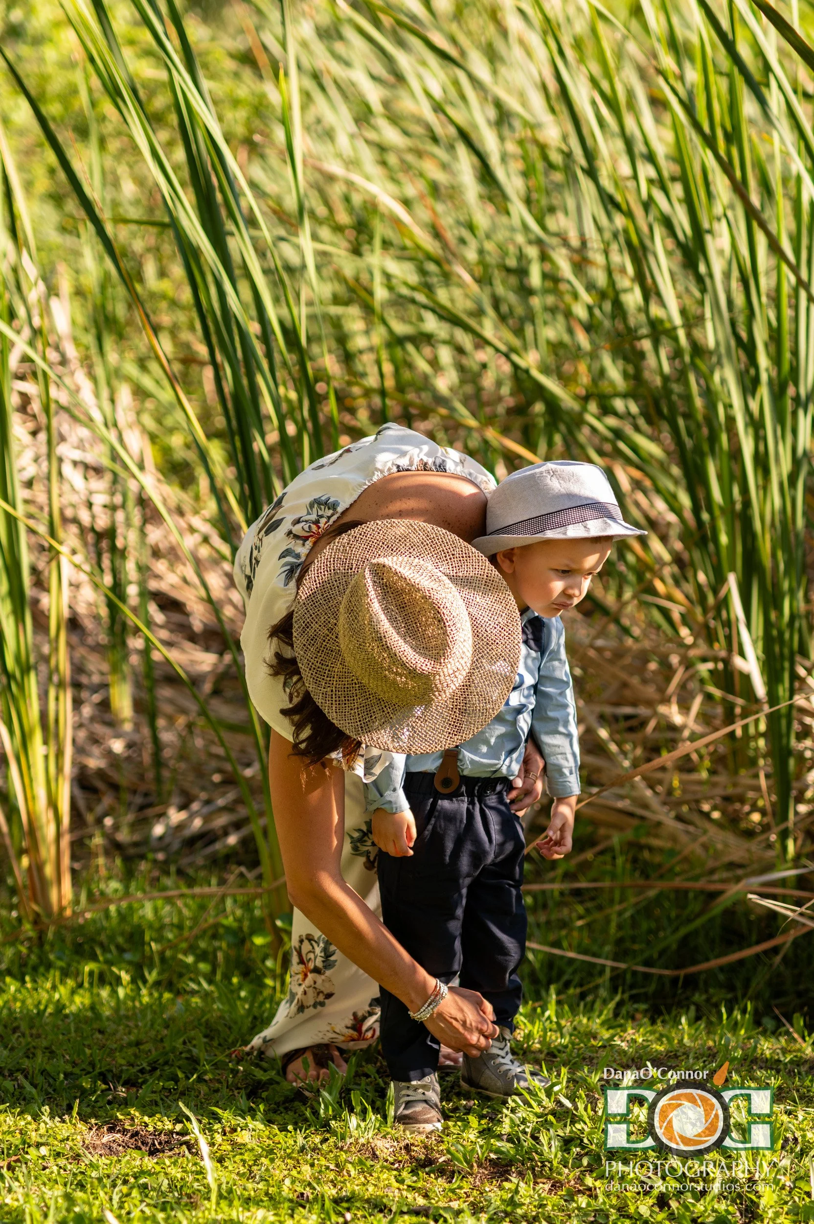 A woman and a child stand in green grass surrounded by tall reeds, with the woman holding the child's shoe while tying it, both wearing sun hats, on a sunny day.