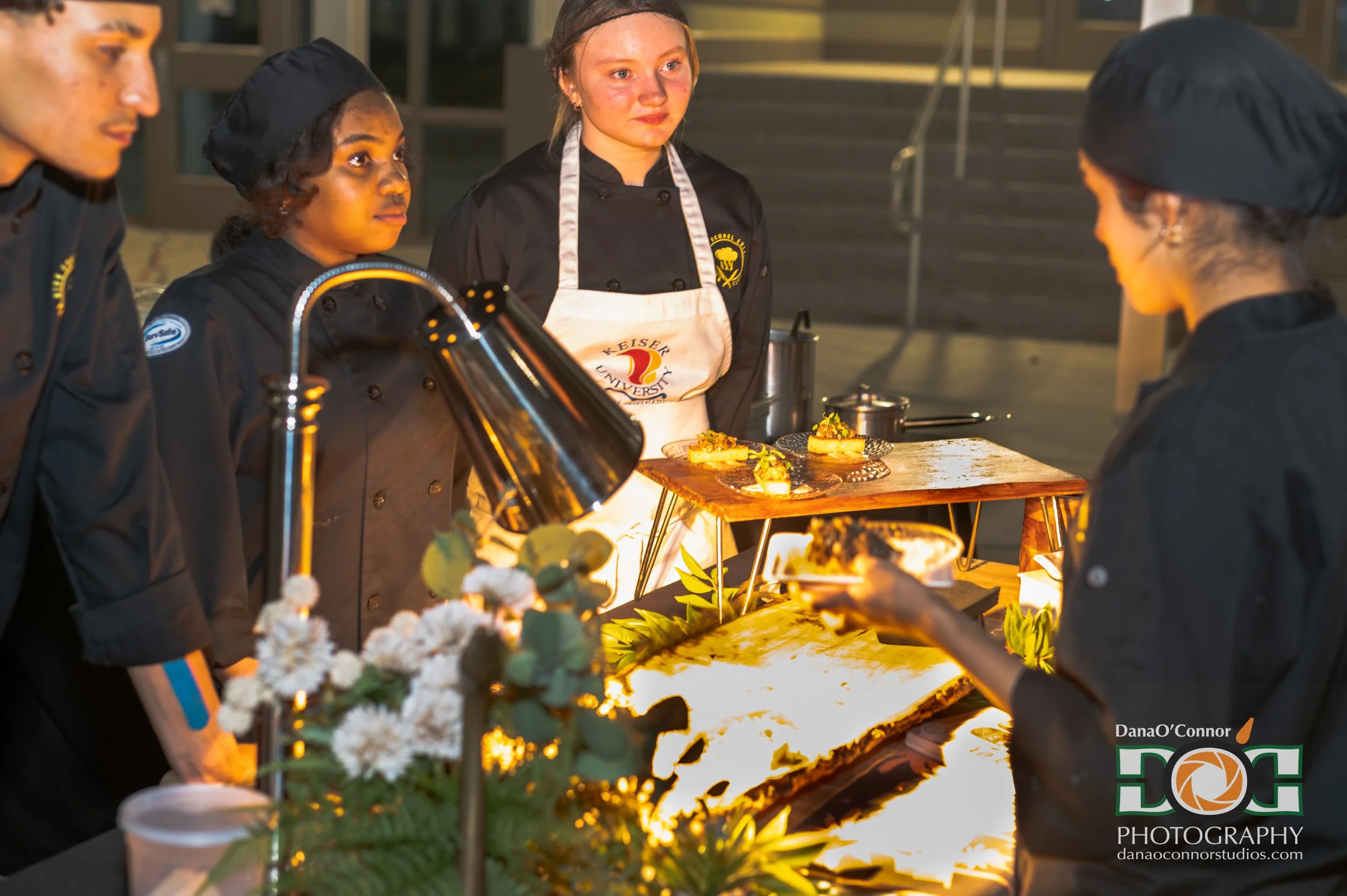 Group of four chefs in black uniforms and aprons preparing food at an outdoor event during evening, with a decorated table, flowers, and warm lighting.
