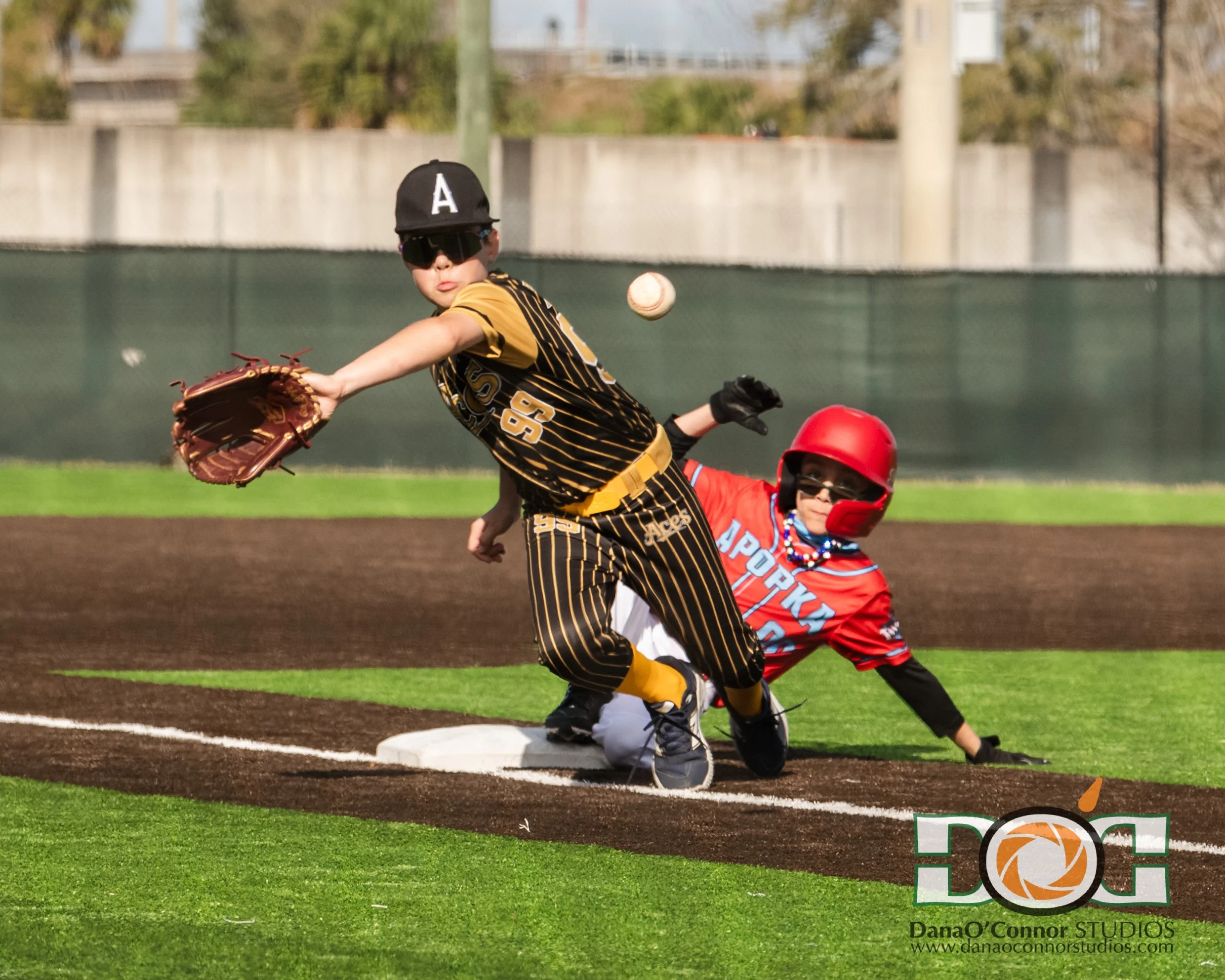 A young baseball player in a yellow and black uniform is reaching to catch a baseball with his glove at first base. Another player in a red helmet and jersey appears to be sliding or falling near the base during a game.